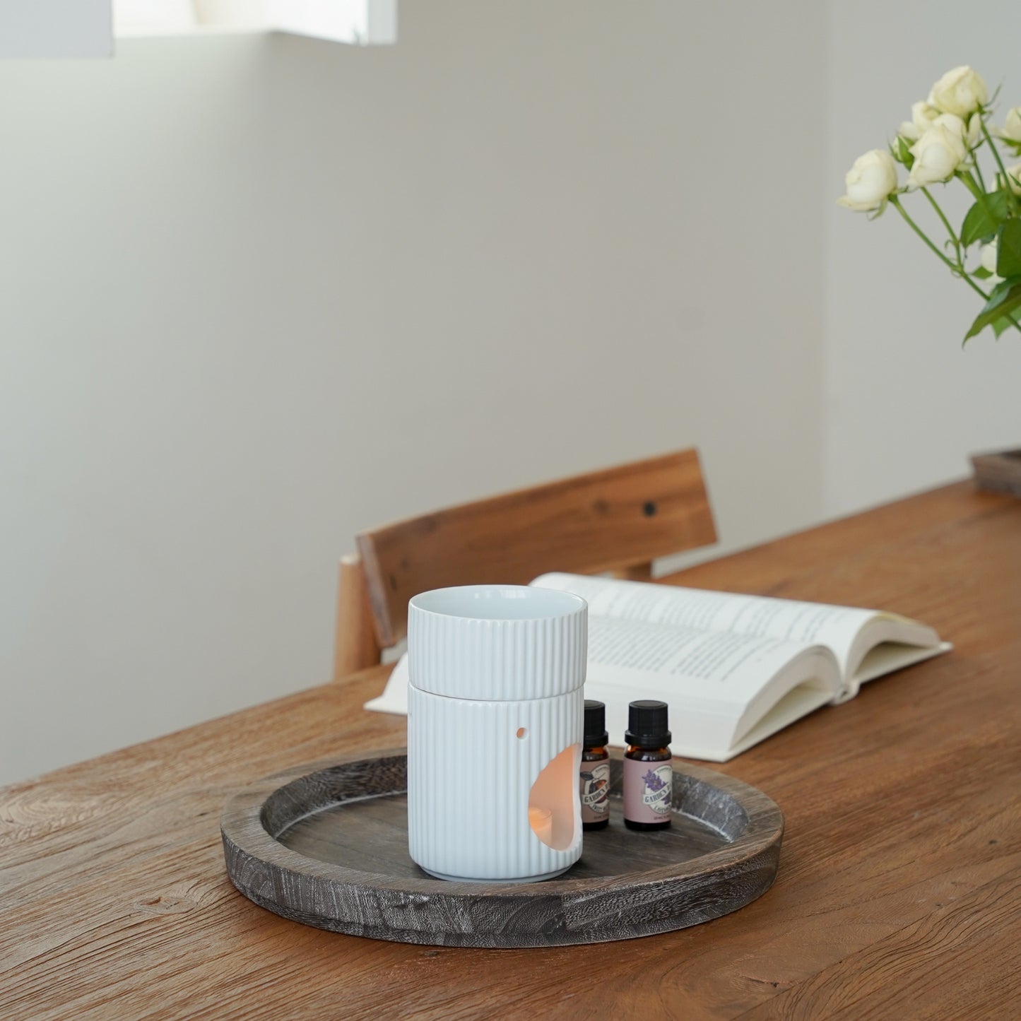 창가 쪽 나무 테이블 위 라운드 트레이에 올려진 세로 골 패턴 화이트 오일버너와 티라이트 캔들, 옆에 가든데이즈 에센셜오일 병과 펼쳐진 책이 보이는 연출 사진