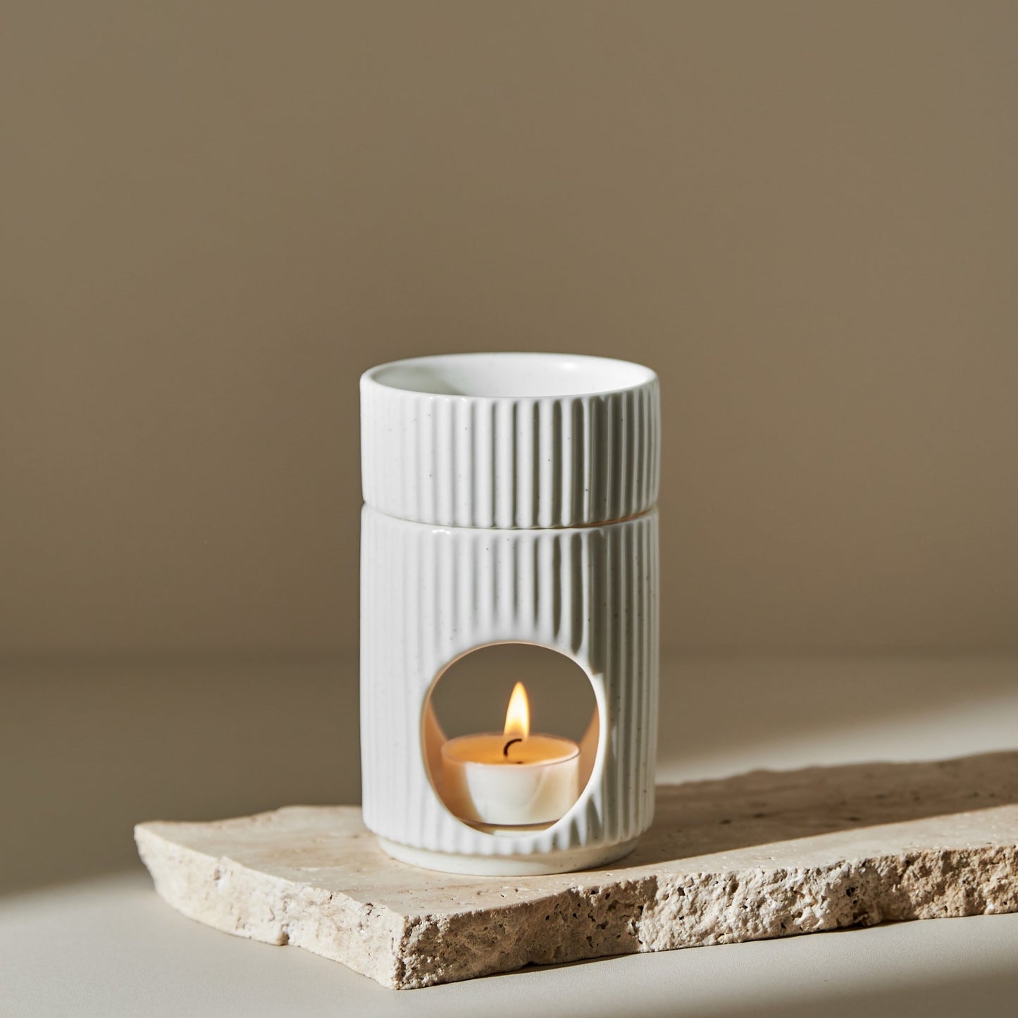 거친 질감의 스톤 받침 위에 세워진 세로 골 패턴의  원통형 오일버너와 안에서 은은하게 켜진 티라이트 캔들이 보이는 제품 연출 사진