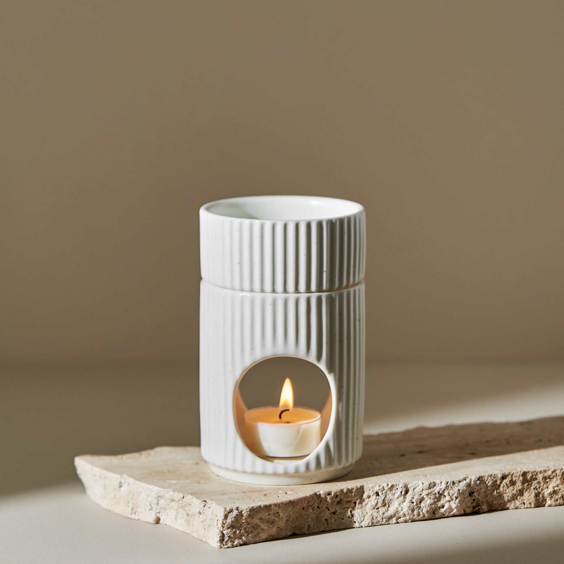 거친 질감의 스톤 받침 위에 세워진 세로 골 패턴의  원통형 오일버너와 안에서 은은하게 켜진 티라이트 캔들이 보이는 제품 연출 사진