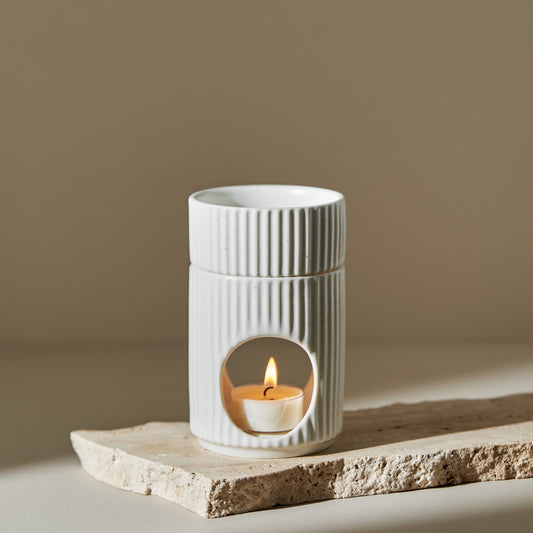 거친 질감의 스톤 받침 위에 세워진 세로 골 패턴의  원통형 오일버너와 안에서 은은하게 켜진 티라이트 캔들이 보이는 제품 연출 사진