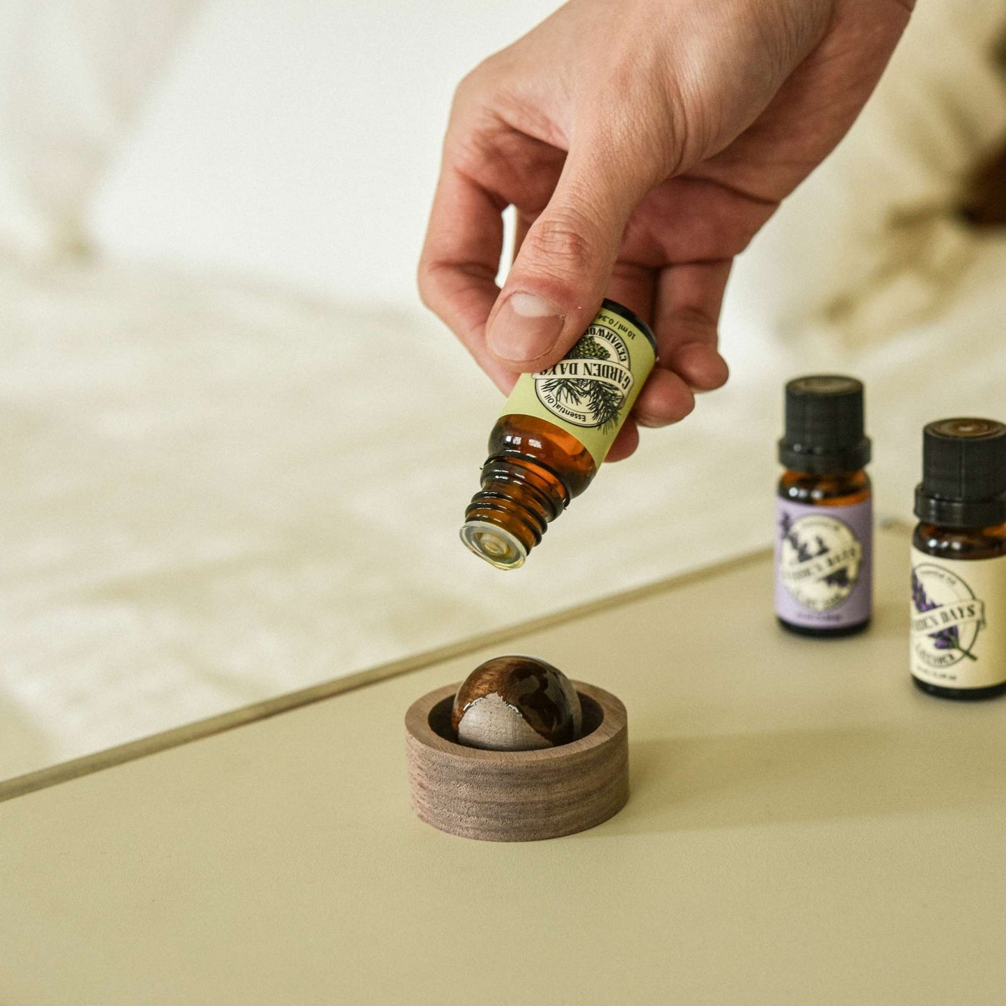 밝은 침구 옆 크림색 상판 위에 놓인 호두 브리즈볼 위로 에센셜오일을 떨어뜨리는 손과 옆에 세워둔 가든데이즈 에센셜오일 병들이 보이는 연출 사진