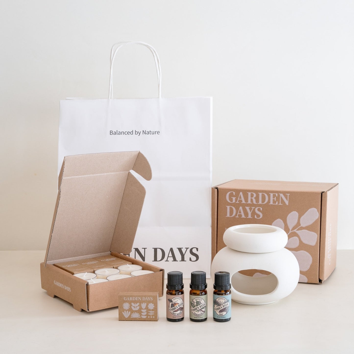 화이트 배경의 테이블 위에 조약돌 도자기 오일버너, 에센셜오일 3종, 소이캔들, 성냥, 그리고 가든데이즈 로고 선물박스와 쇼핑백이 모두 펼쳐져 있는 조약돌 오일버너 선물세트 구성 사진