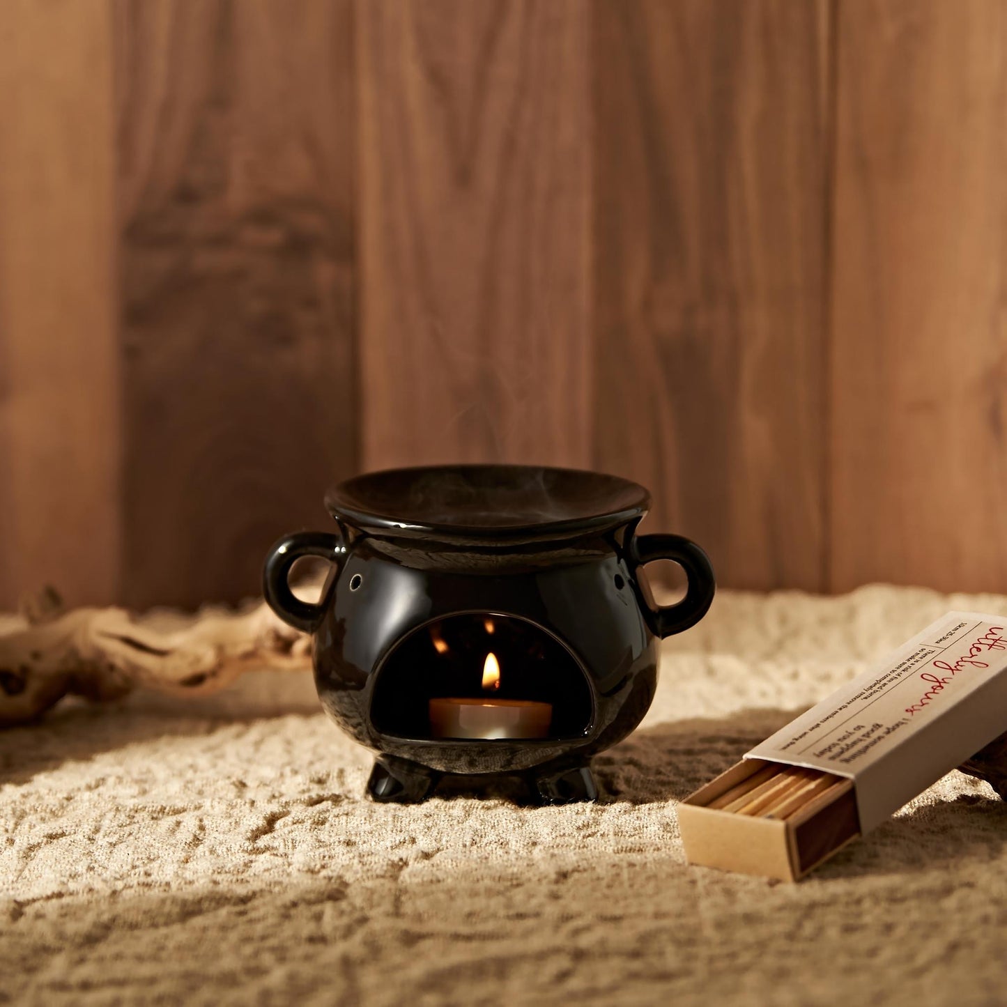 우드 배경과 러그 위에 단독으로 놓인 블랙 가마솥 모양 오일버너와 은은한 불빛의 티라이트 캔들, 옆에 성냥갑이 보이는 제품 연출 사진