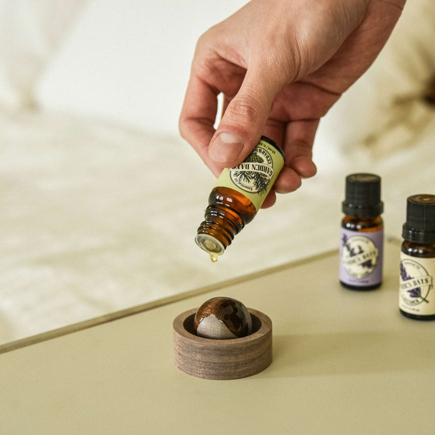 침대 옆 상판 위 호두 브리즈볼 우드 발향 제품 위로 가든데이즈 천연 100% 에센셜오일을 떨어뜨리는 손과 옆에 세워진 오일 병들이 보이는 사진