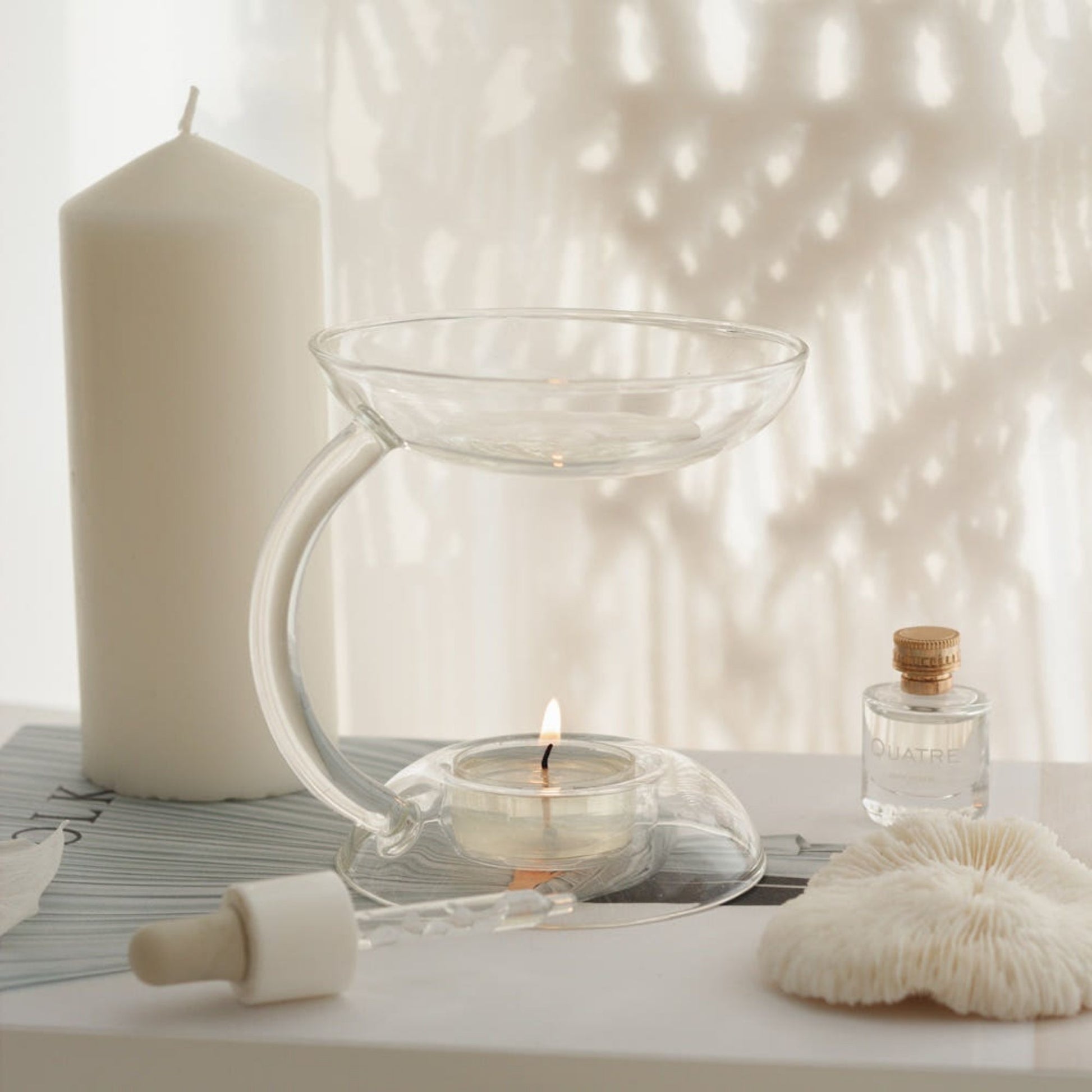 빛과 그림자가 드리운 창가 책 더미 위에 놓인 투명한 유리 오일버너와 안에서 켜진 티라이트 캔들, 옆의 화이트 기둥 캔들과 장식이 함께 보이는 연출 사진