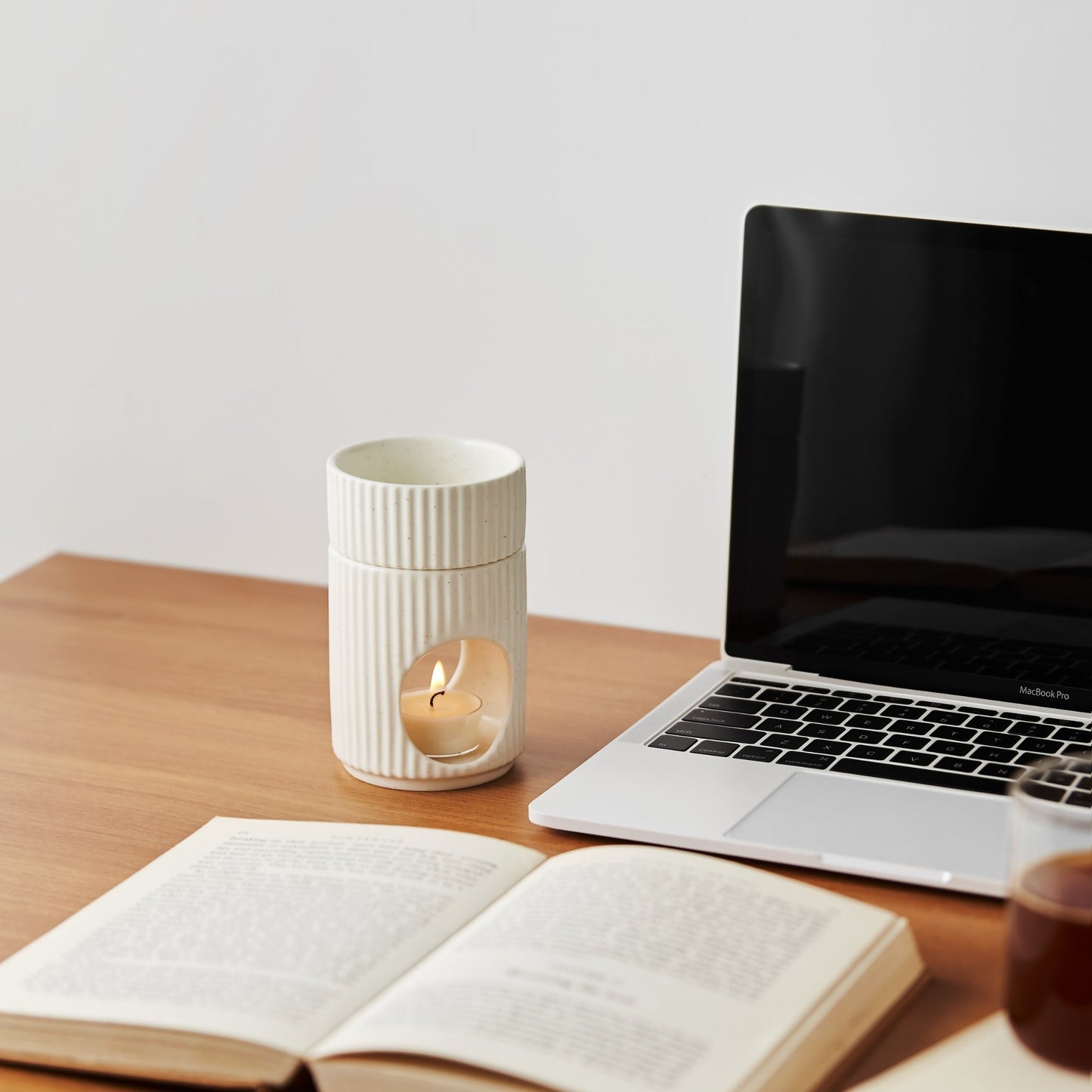노트북과 펼쳐진 책이 놓인 책상 위에 세워진 세로 골 패턴 도자기 오일버너와 따뜻한 불빛의 티라이트 캔들이 보이는 작업 공간 연출 사진
