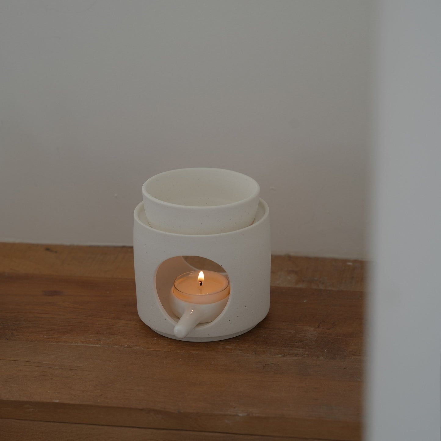 나무 선반 위 벽 쪽에 놓인 화이트 세라믹 오일버너와 안에서 켜진 티라이트 캔들이 보이는 사진