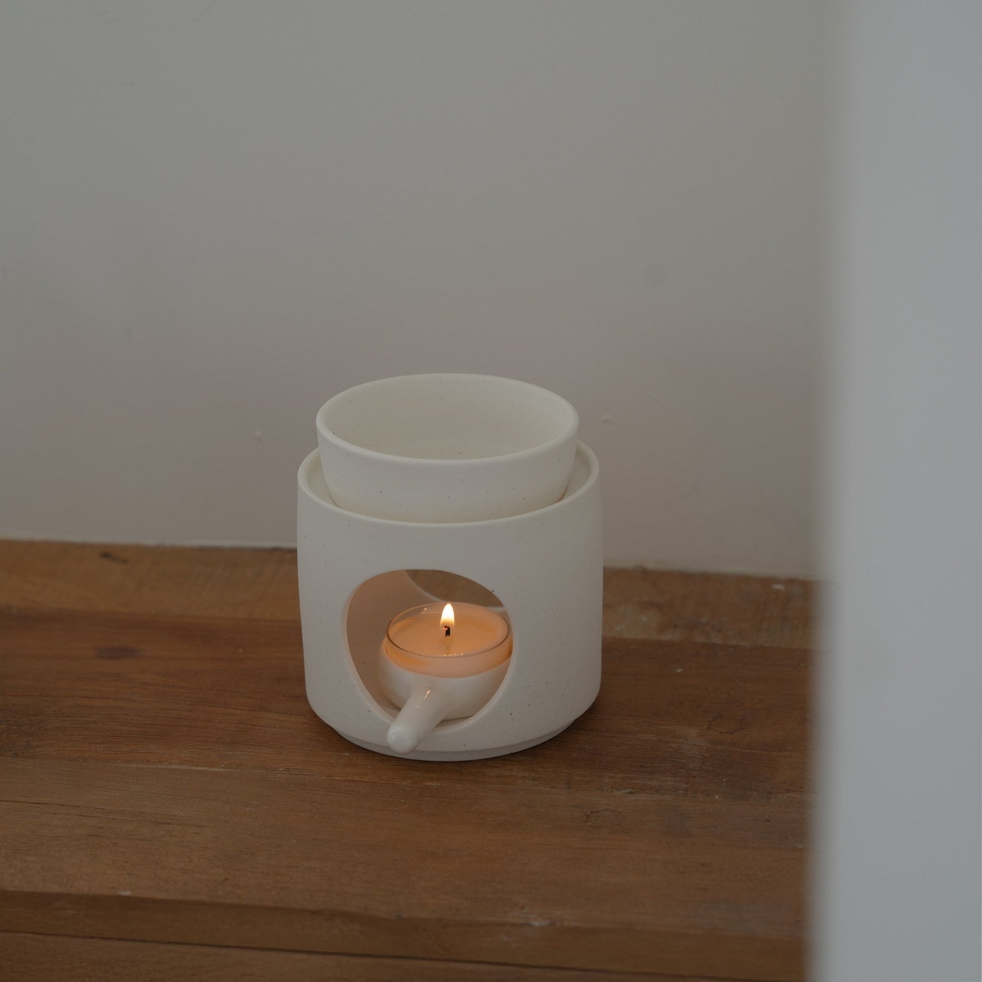 나무 선반 위 벽 쪽에 놓인 화이트 세라믹 오일버너와 안에서 켜진 티라이트 캔들이 보이는 사진