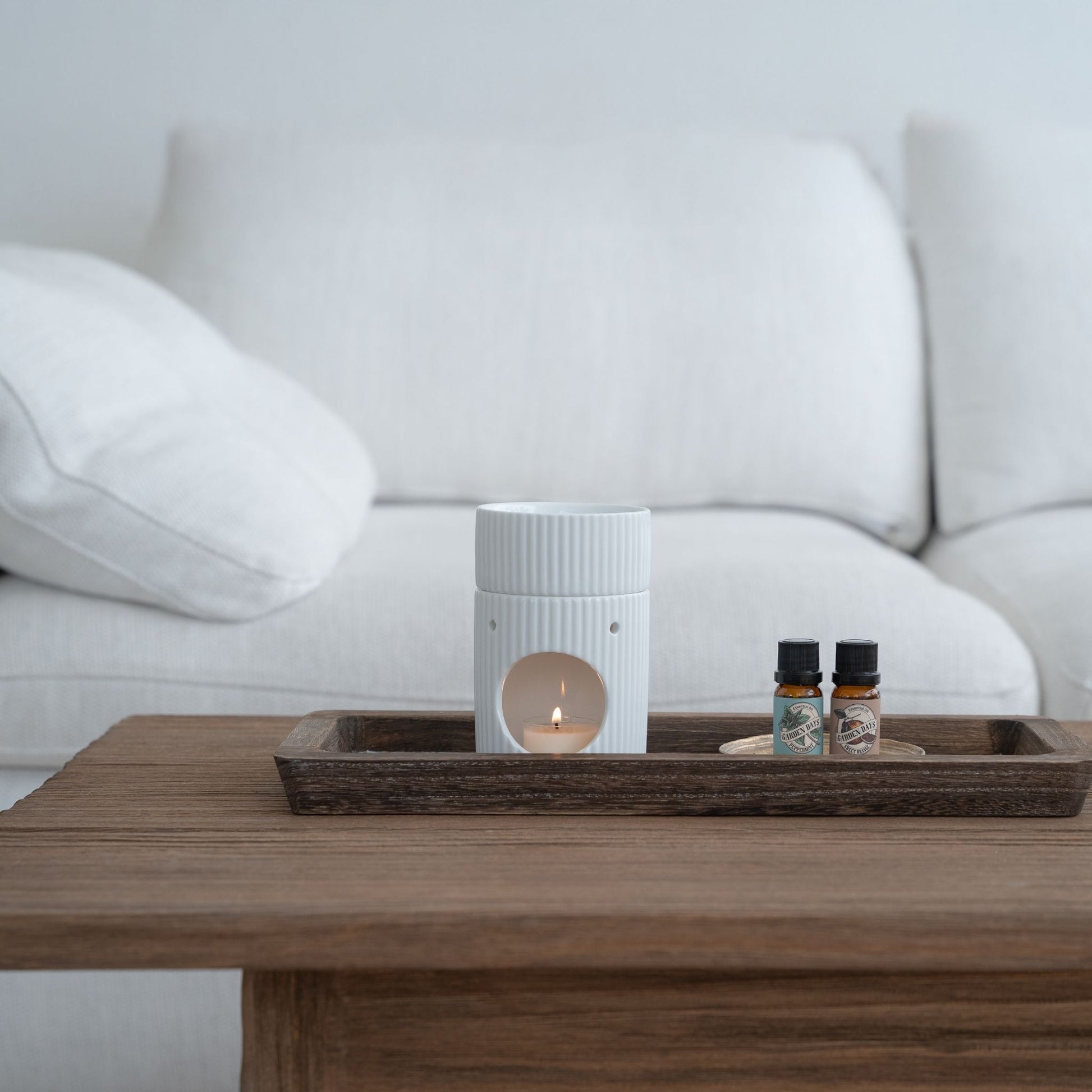 화이트 소파 앞 나무 테이블 위 트레이에 라운드 오일버너와 가든데이즈 에센셜오일 2병이 함께 놓인 거실 연출 사진