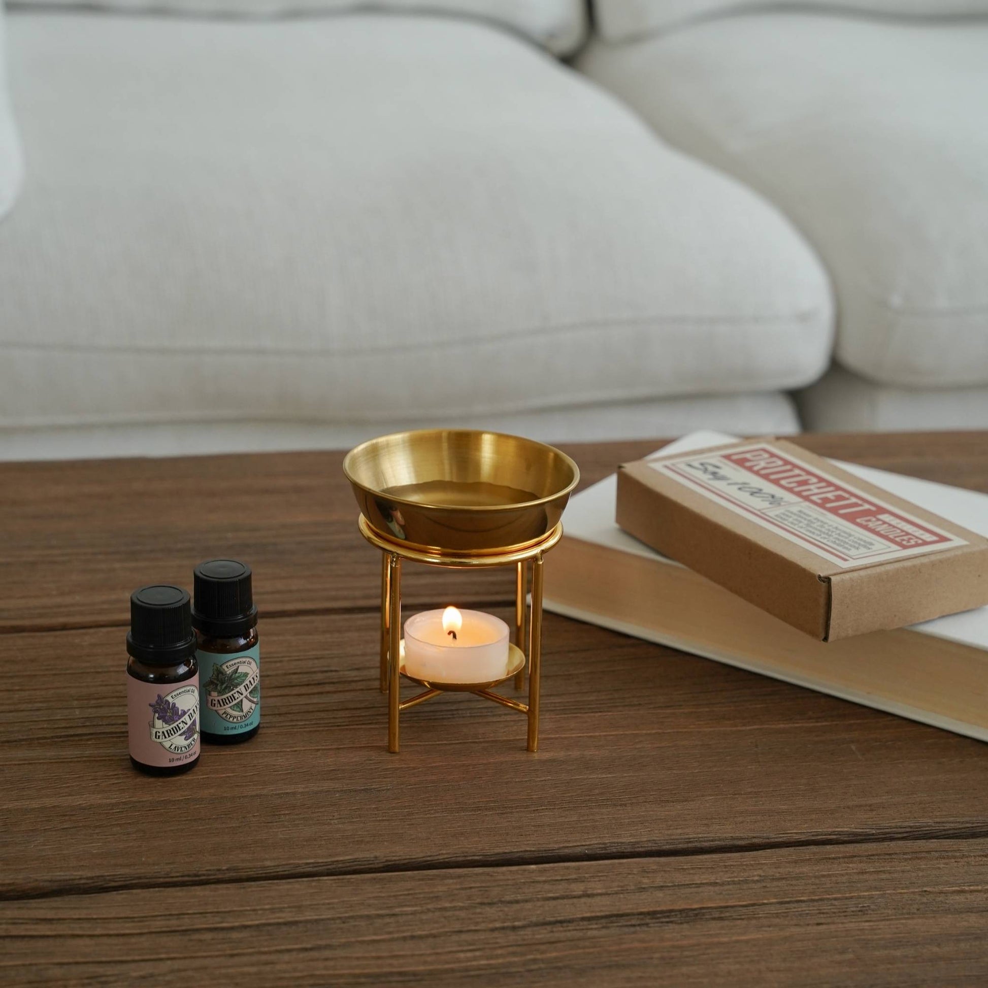 소파 앞 나무 테이블 위 골드 스탠드 오일버너와 불이 켜진 티라이트 캔들, 가든데이즈 에센셜오일 병이 놓인 거실 연출 사진