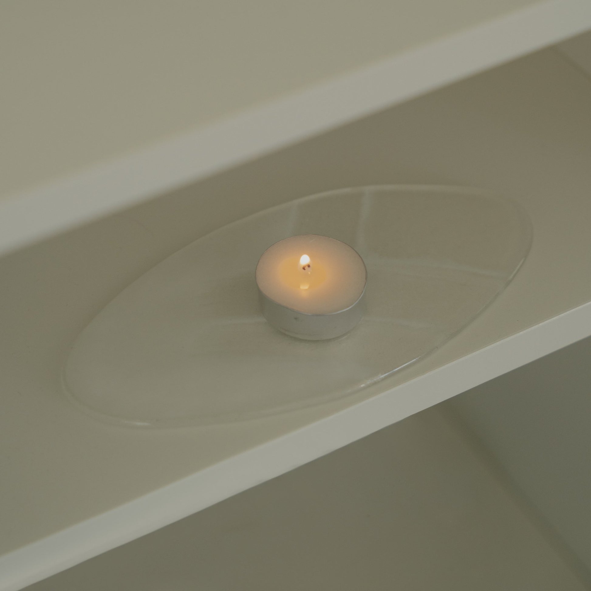 화이트 선반 위 투명한 받침 접시 위에 놓인 하나의 티라이트 캔들이 보이는 연출 사진