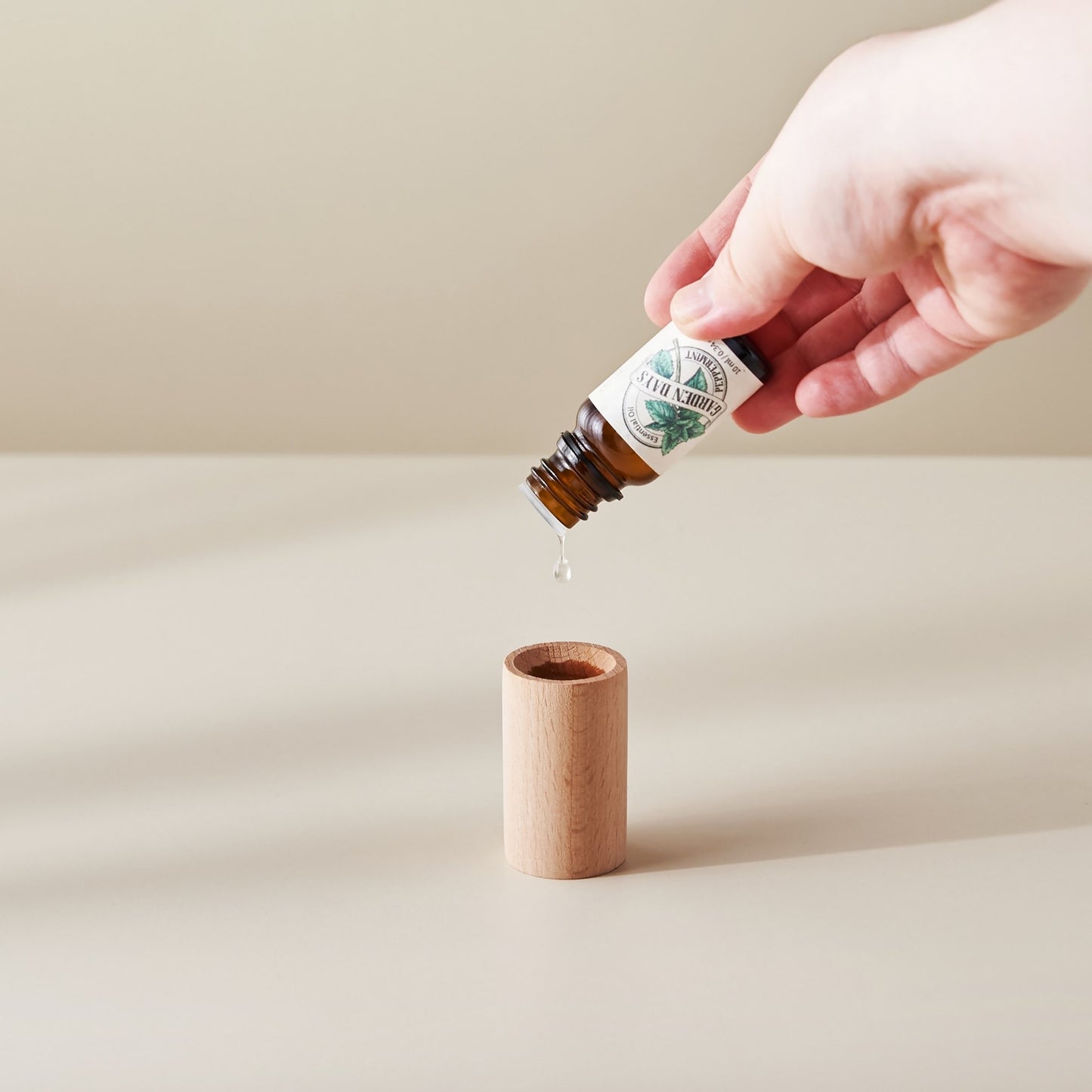 밝은 배경 위에 세워진 너도밤나무 원목 실린더 발향 우드 위로 에센셜오일을 떨어뜨리는 손이 보이는 사용 방법 연출 사진