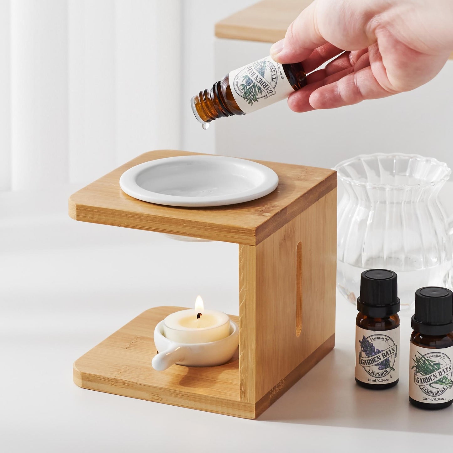 밝은 테이블 위 우드 오일버너와 불이 켜진 티라이트 캔들, 옆에 가든데이즈 에센셜오일 병 두 개와 에센셜오일을 사용하는 연출 사진