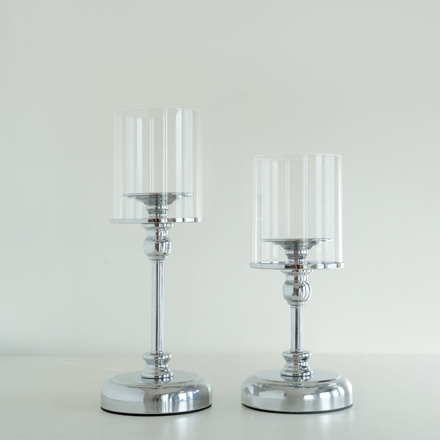 밝은 배경 앞에 실버 베이스와 투명 유리 갓이 있는 스탠드 캔들 홀더 두 개를 나란히 세운 제품 단독 사진