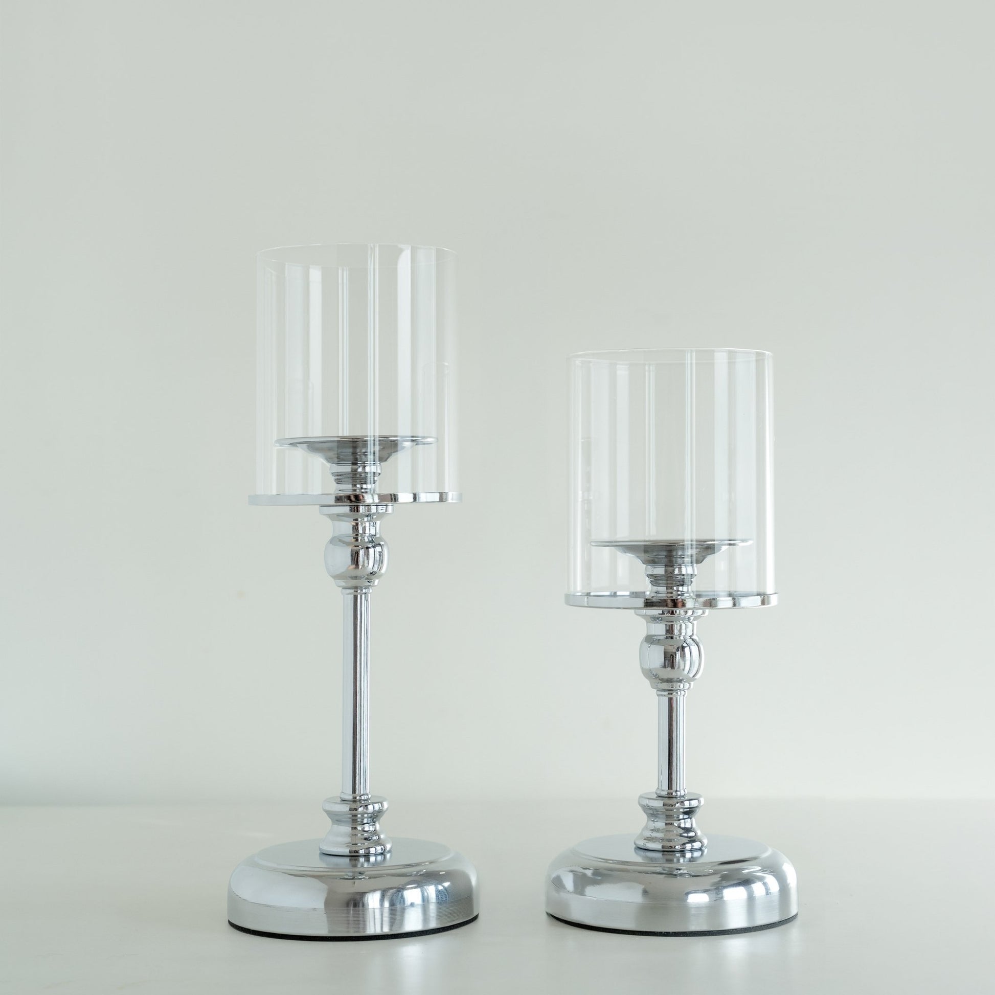 밝은 배경 앞에 실버 베이스와 투명 유리 갓이 있는 스탠드 캔들 홀더 두 개를 나란히 세운 제품 단독 사진