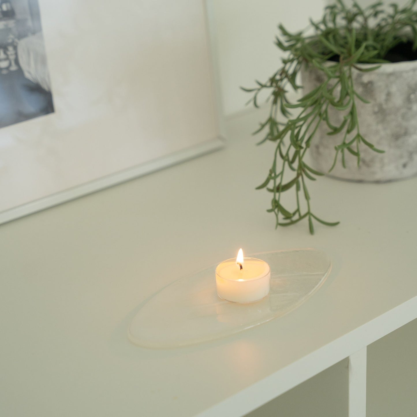 화이트 선반 위 투명 받침 접시 위에 하나만 올려둔 소이 티라이트 캔들이 은은하게 켜져 있는 연출 사진