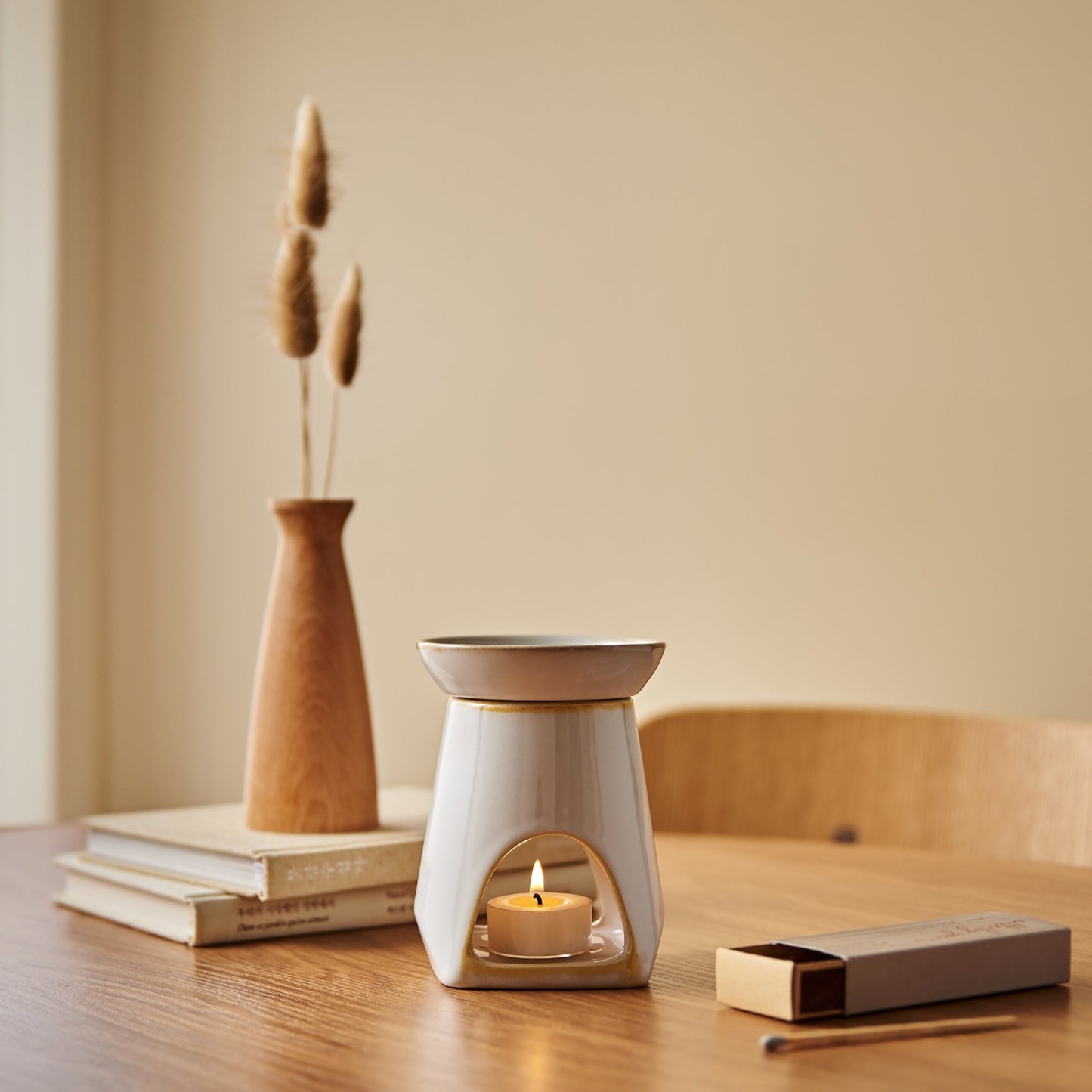 나무 테이블 위 책 더미와 우드 베이스 꽃병 옆에 놓인 화이트 세라믹 옐로우 오일버너와 불이 켜진 티라이트 캔들이 보이는 연출 사진