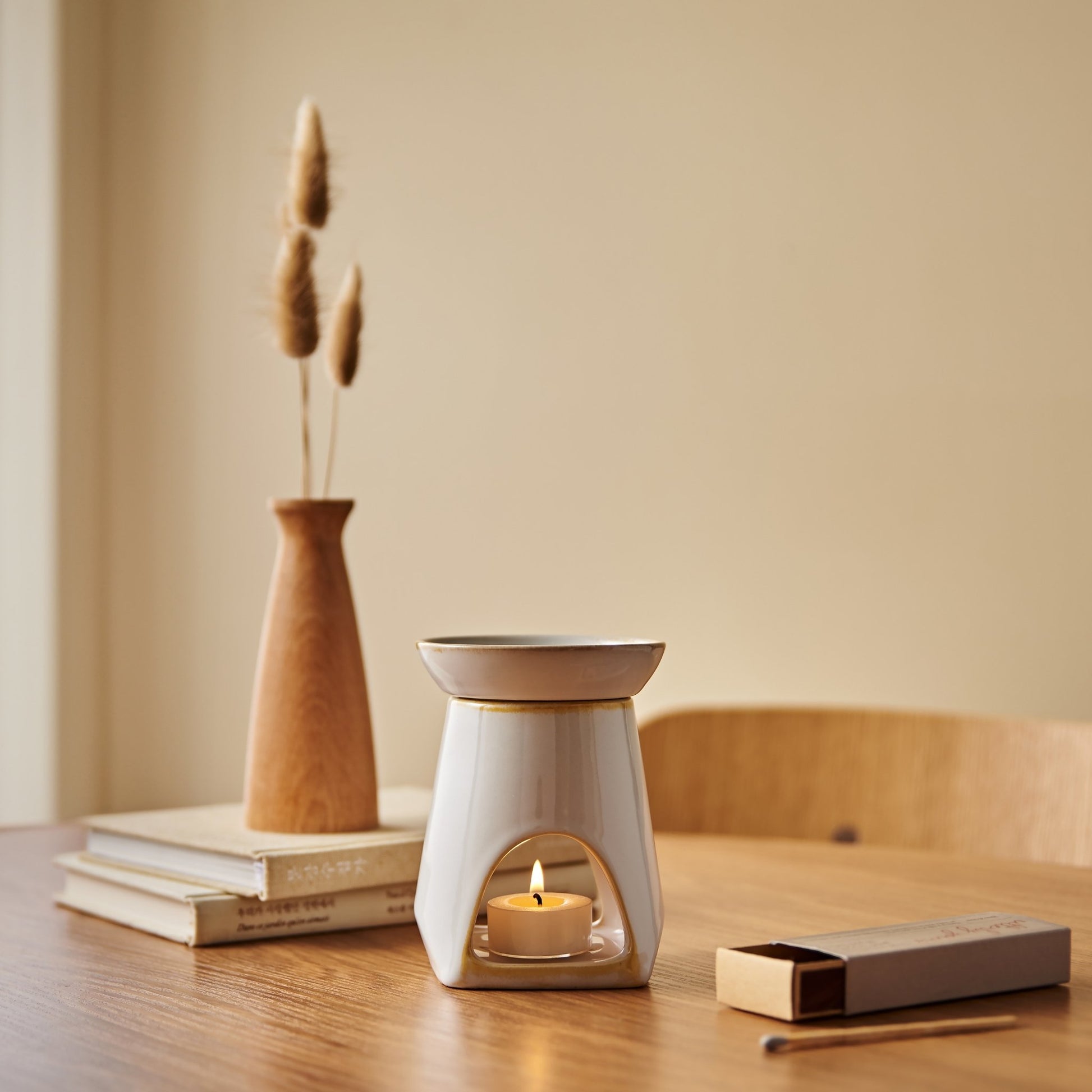 나무 테이블 위 책 더미와 우드 베이스 꽃병 옆에 놓인 화이트 세라믹 옐로우 오일버너와 불이 켜진 티라이트 캔들이 보이는 연출 사진