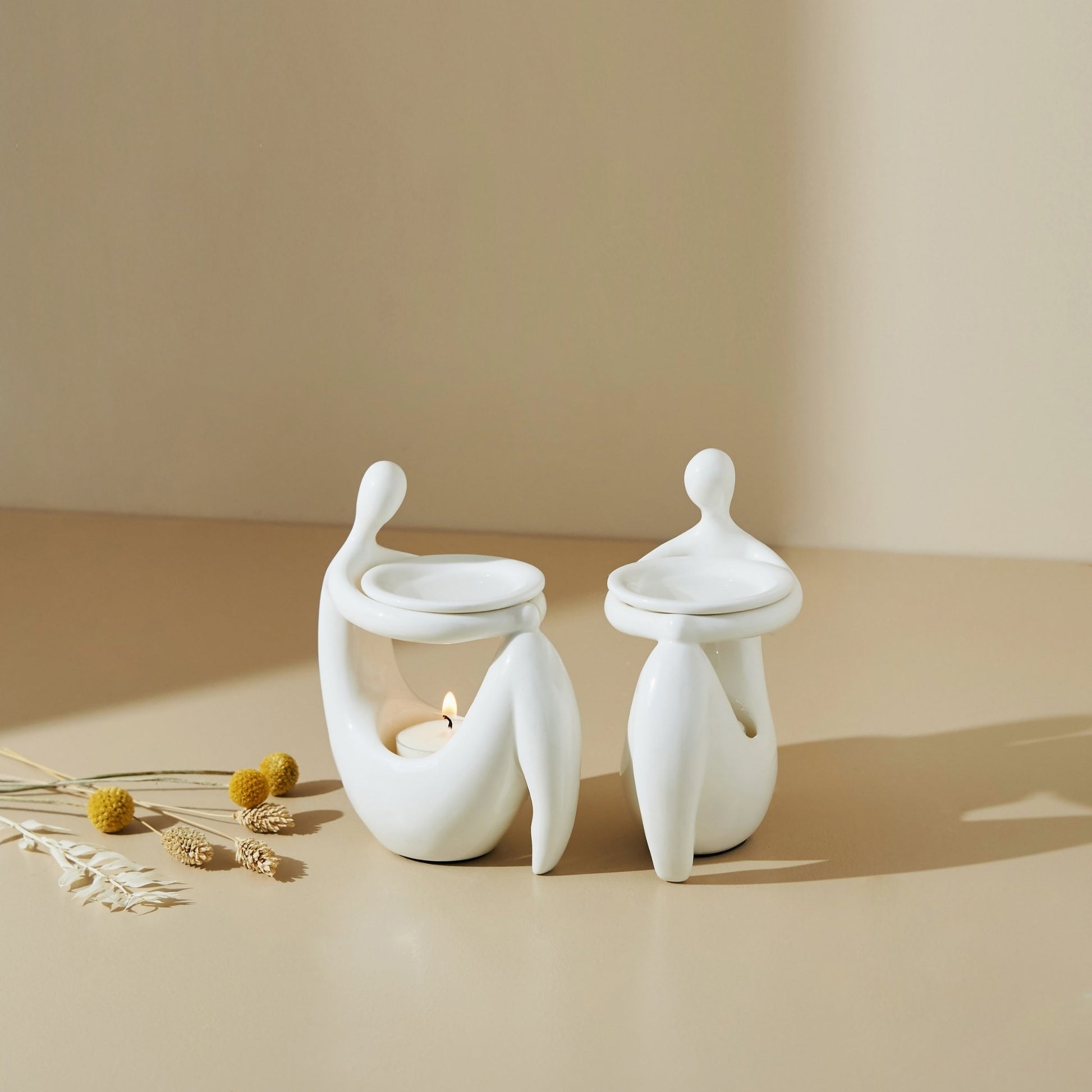 베이지 톤 배경 위에 단독으로 놓인 화이트 조형적인 세라믹 오일버너와 불이 켜진 티라이트 캔들, 주변에 흩뿌려진 작은 꽃과 허브가 보이는 제품 연출 사진