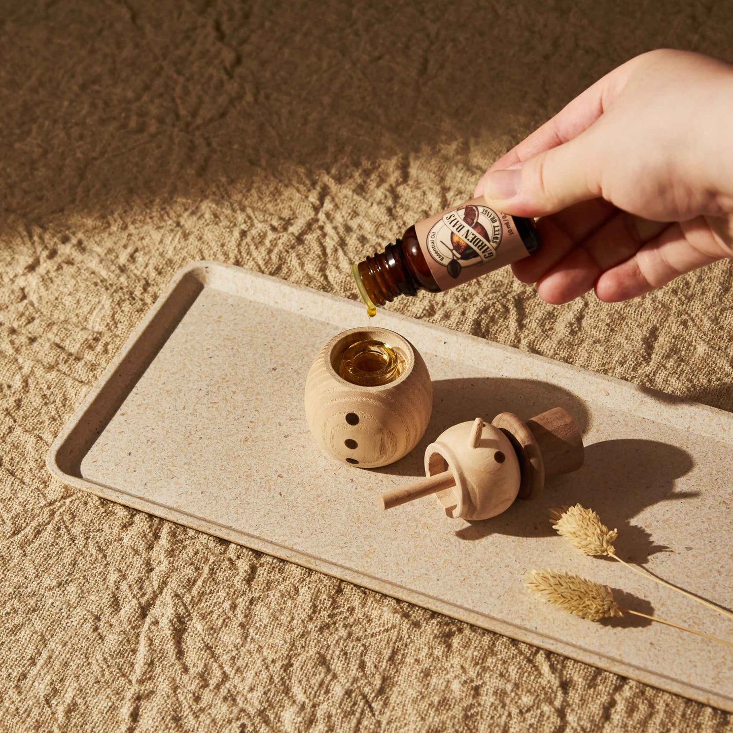 트레이 위에 뚜껑을 분리해 둔 우드 스노우맨 디퓨저와 내부 유리병 위로 에센셜오일을 떨어뜨리는 손이 보이는 사용 방법 연출 사진