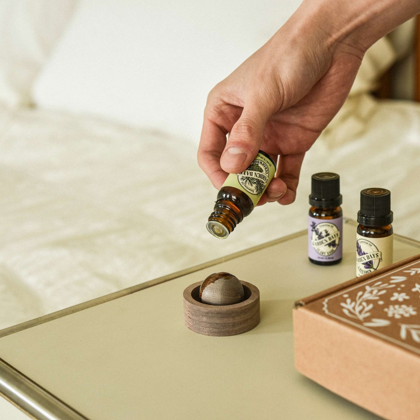 침대 옆 테이블 위에 놓인 호두 브리즈볼 위로 에센셜오일을 떨어뜨리는 손과 옆에 세워진 에센셜오일 병들이 보이는 침실 사용 연출 사진