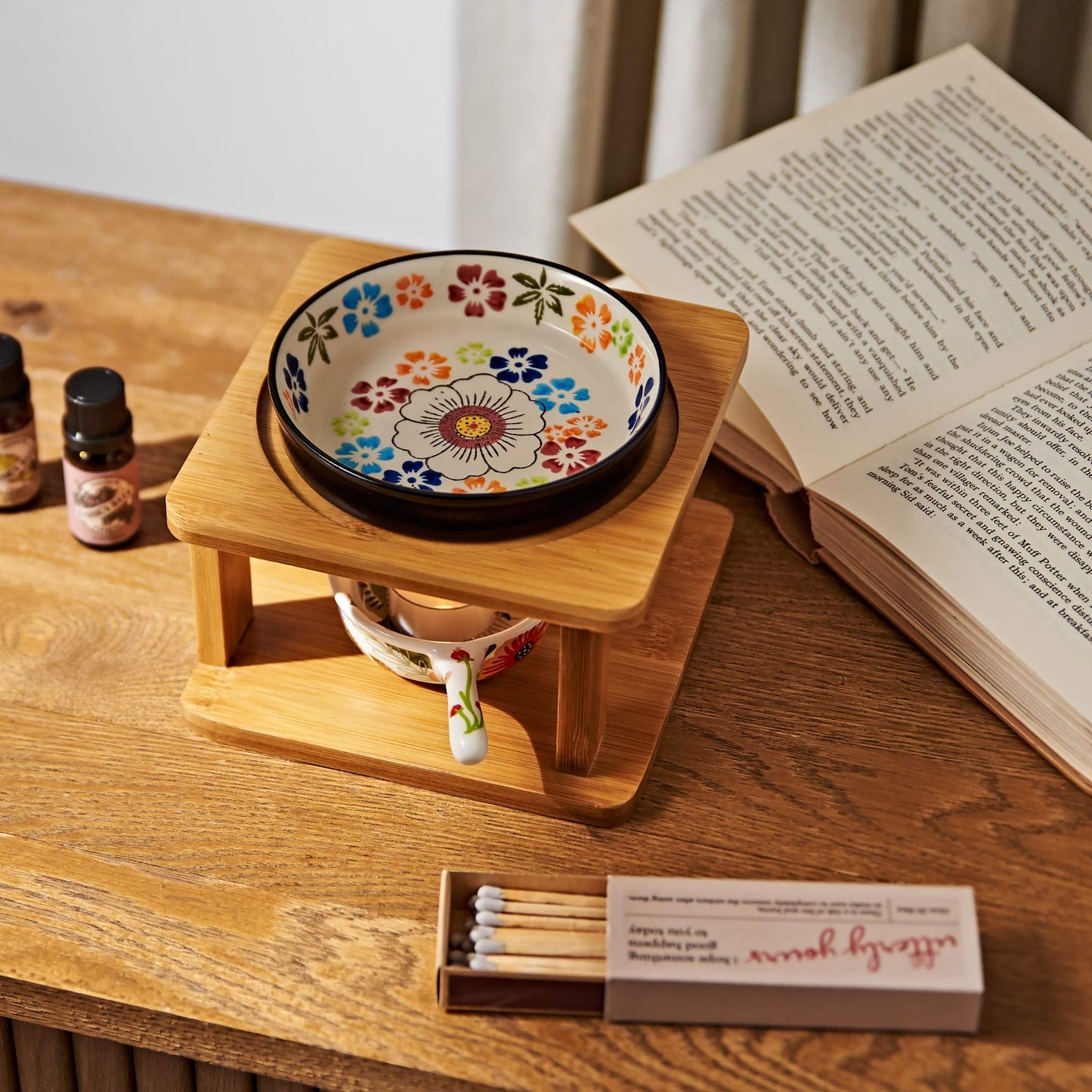 나무 테이블 위 펼쳐진 책과 성냥갑 옆에 놓인 오일버너, 위에 올려진 플라워 패턴 세라믹 볼과 아래 불이 켜진 티라이트 캔들, 가든데이즈 에센셜오일 병이 함께 보이는 연출 사진