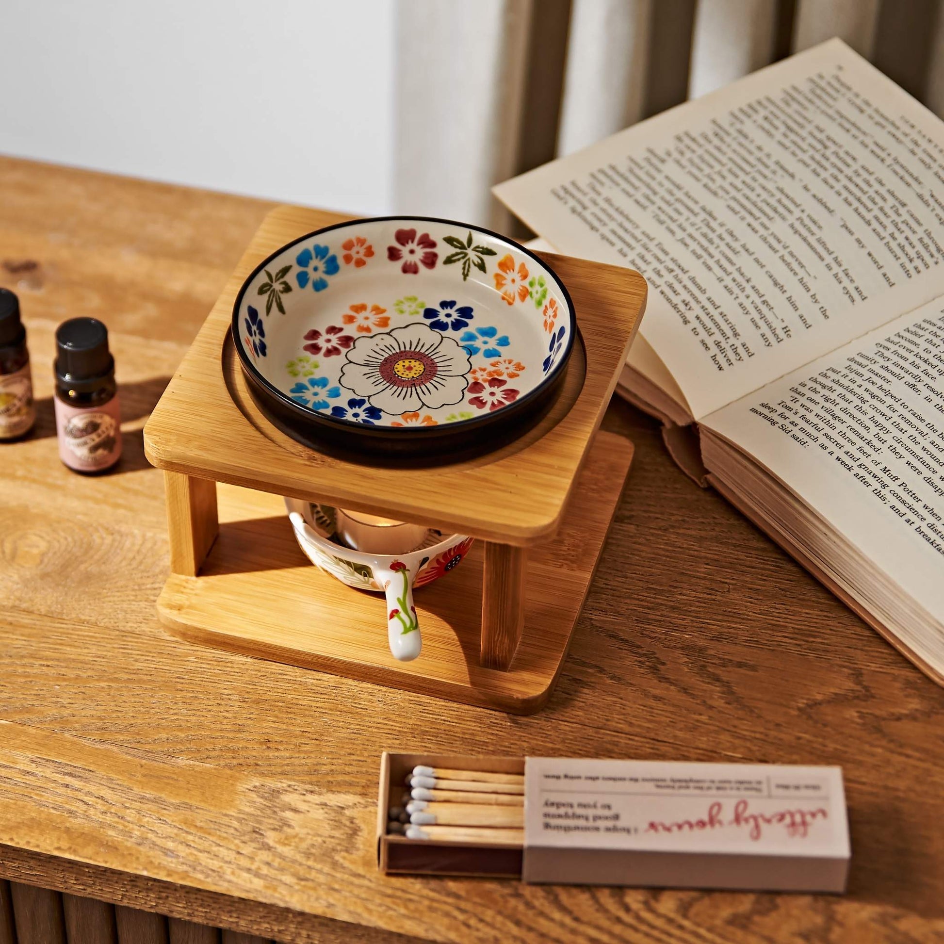 나무 테이블 위 펼쳐진 책과 성냥갑 옆에 놓인 오일버너, 위에 올려진 플라워 패턴 세라믹 볼과 아래 불이 켜진 티라이트 캔들, 가든데이즈 에센셜오일 병이 함께 보이는 연출 사진