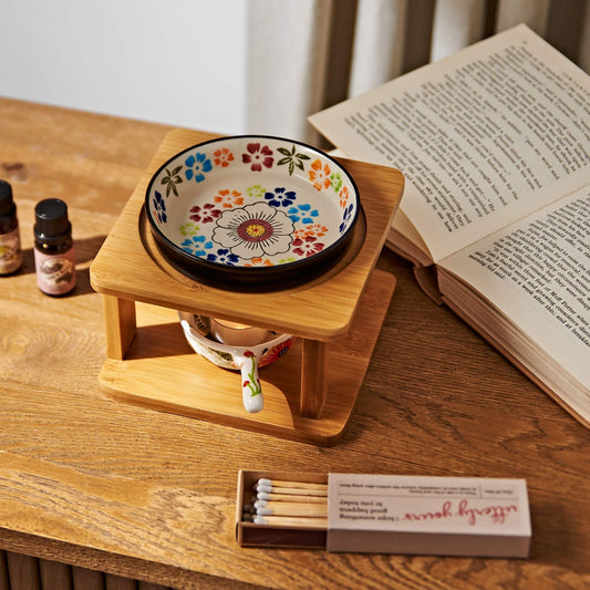 나무 테이블 위 펼쳐진 책과 성냥갑 옆에 놓인 오일버너, 위에 올려진 플라워 패턴 세라믹 볼과 아래 불이 켜진 티라이트 캔들, 가든데이즈 에센셜오일 병이 함께 보이는 연출 사진