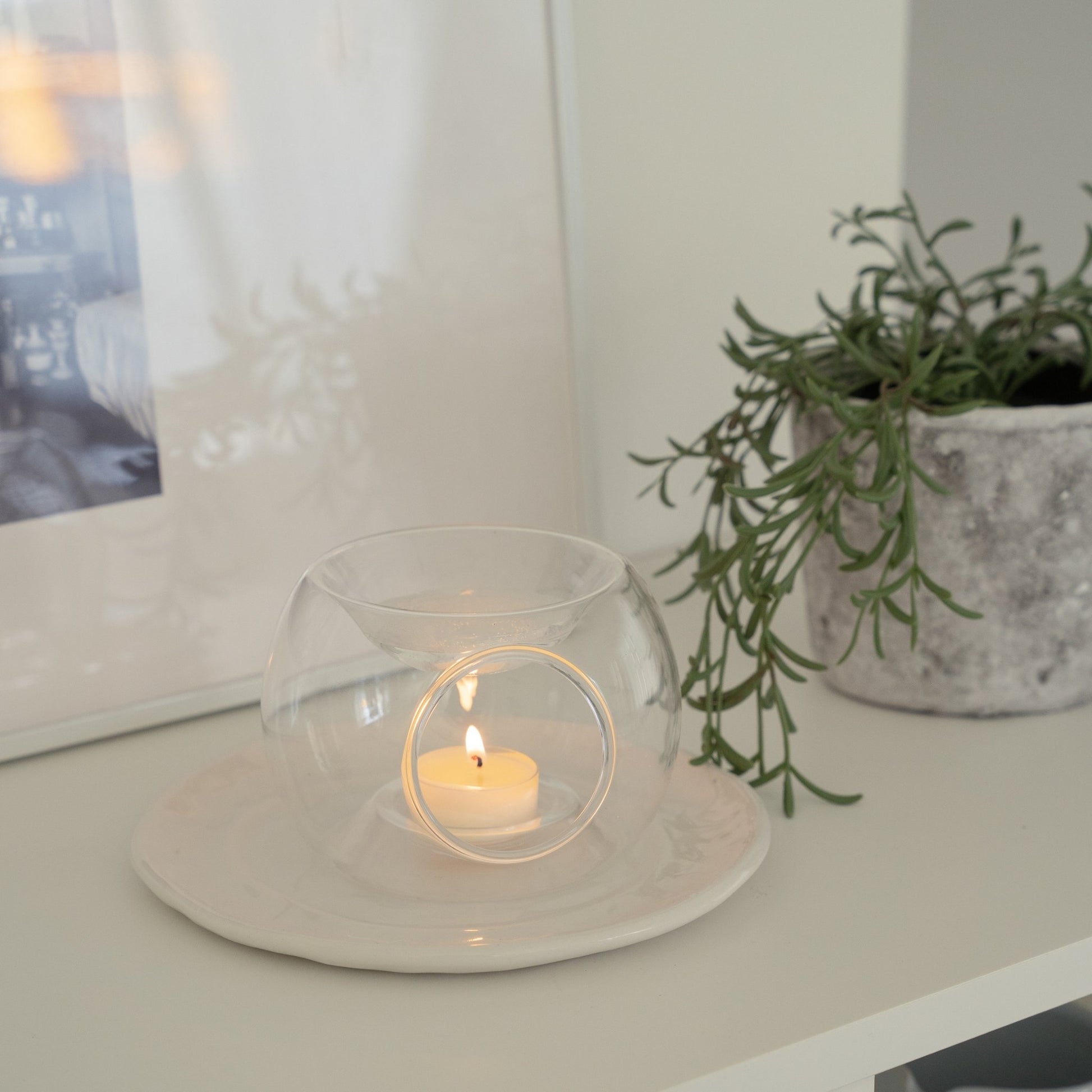 화이트 선반 위 원형 접시 위에 올려진 투명 유리 티라이트 홀더와 안에서 따뜻하게 빛나는 티라이트 캔들, 옆에 액자와 화분이 보이는 연출 사진