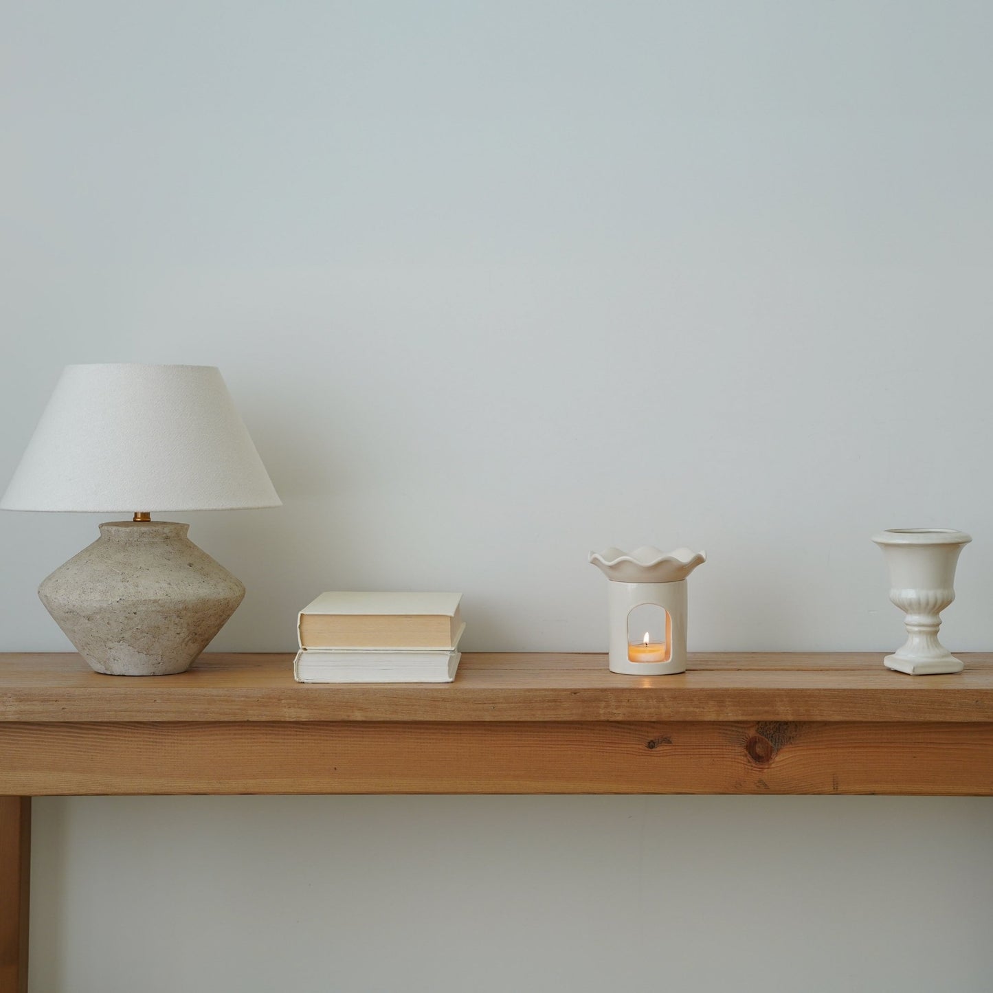 테이블 스탠드와 책들이 놓인 긴 나무 선반 위 한쪽에 배치된 웨이브  모양 화이트 오일버너와 은은한 불빛의 티라이트 캔들이 보이는 인테리어 연출 사진