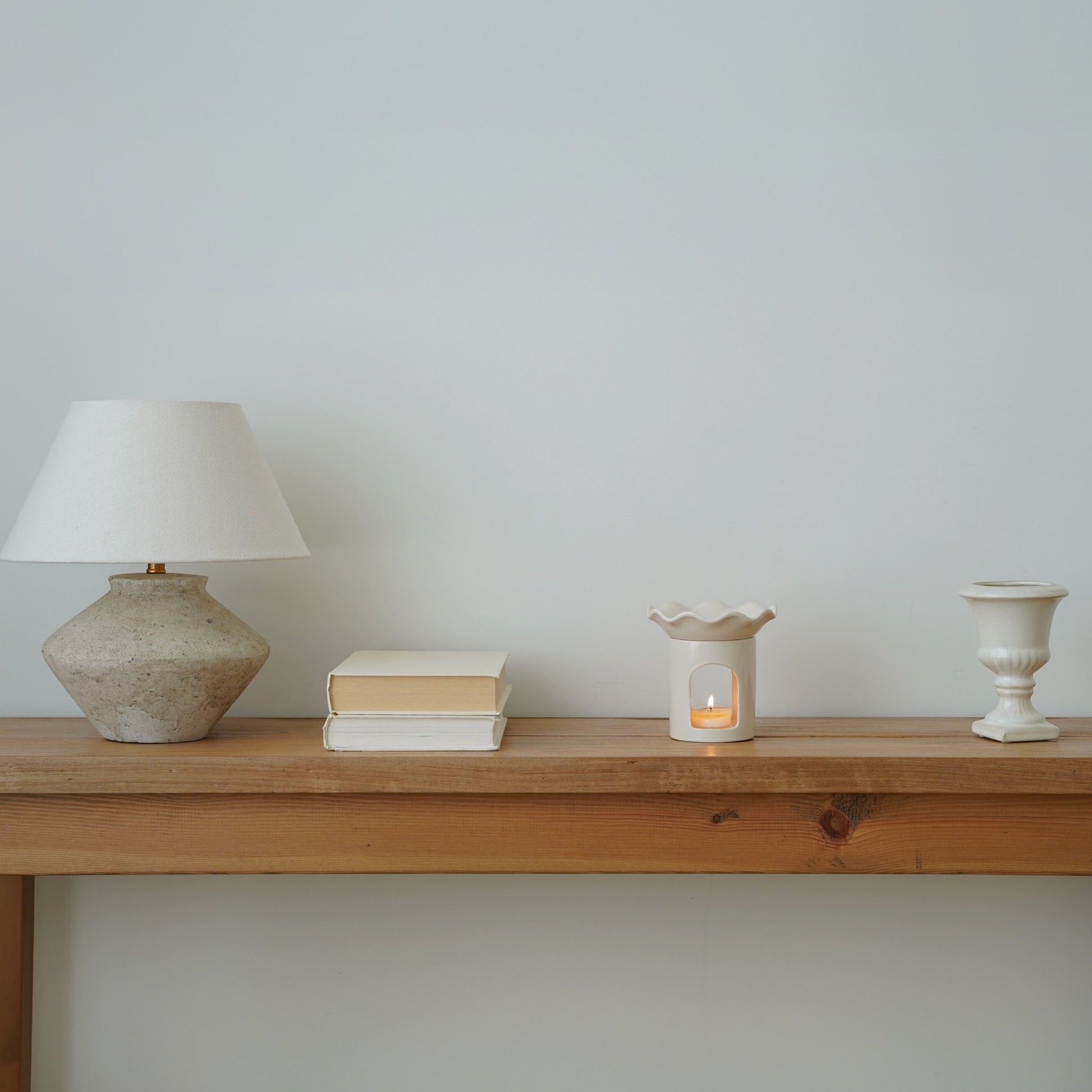 테이블 스탠드와 책들이 놓인 긴 나무 선반 위 한쪽에 배치된 웨이브  모양 화이트 오일버너와 은은한 불빛의 티라이트 캔들이 보이는 인테리어 연출 사진
