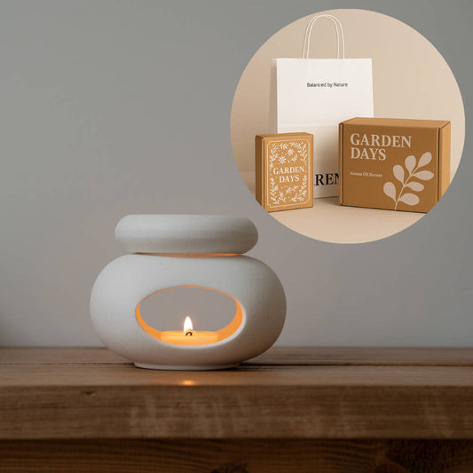 나무 테이블 위에서 티라이트 캔들이 켜진 하얀 조약돌 도자기 오일버너가 보이고, 상단 원형 인셋 이미지에는 쇼핑백과 선물박스, 에센셜오일·소이캔들·성냥이 포함된 조약돌 오일버너 선물세트 패키지가 함께 보이는 사진