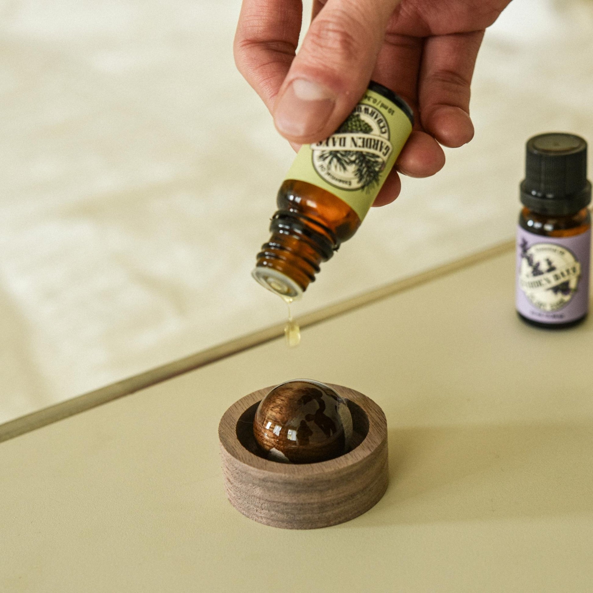 밝은 상판 위에 놓인 호두 브리즈볼 옆으로 가든데이즈 에센셜오일 병 두 개가 세워져 있고, 위에서 에센셜오일을 떨어뜨리는 손이 보이는 클로즈업 사진