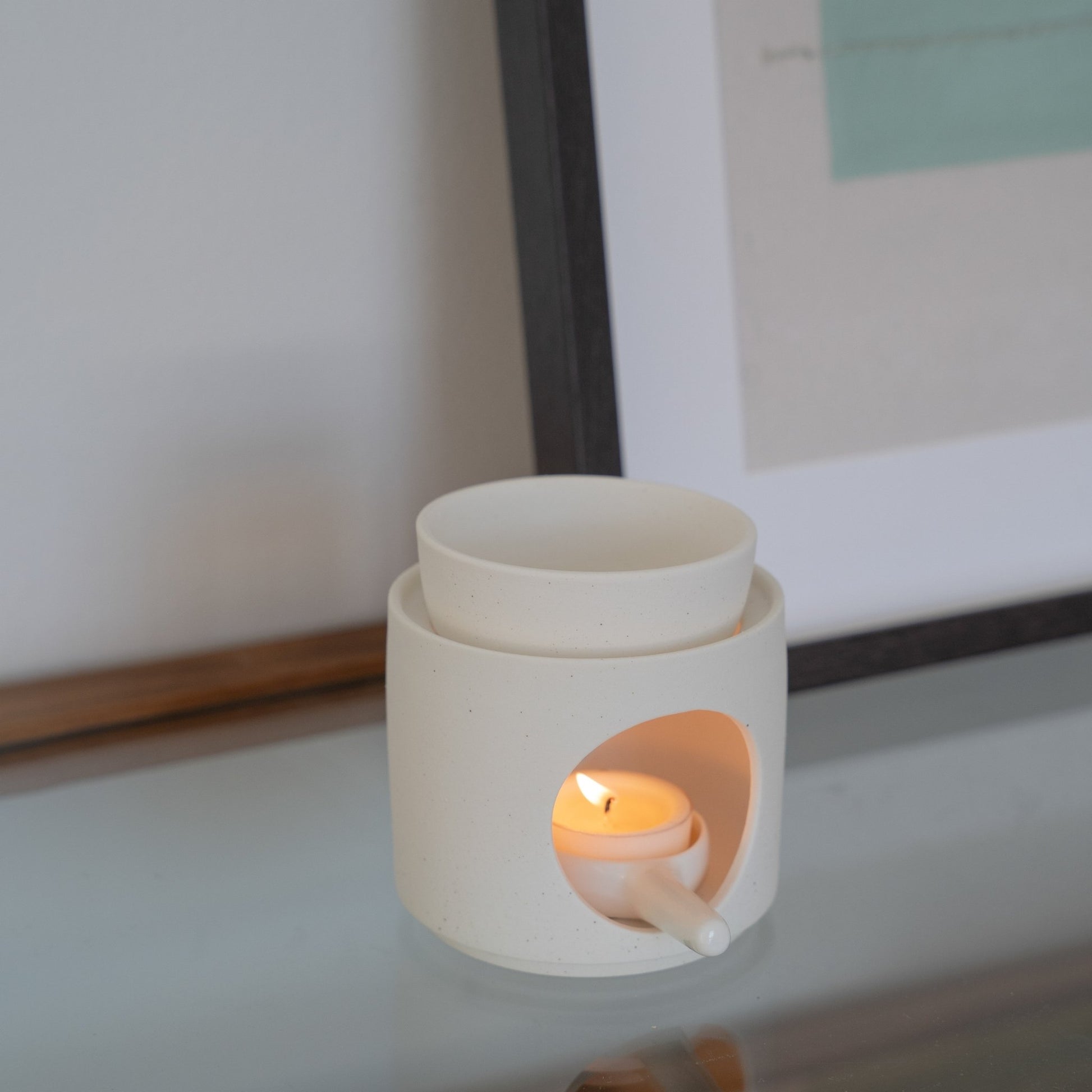 유리 상판 위 액자와 함께 놓인 화이트 세라믹 오일버너와 불이 켜진 티라이트 캔들 클로즈업 사진