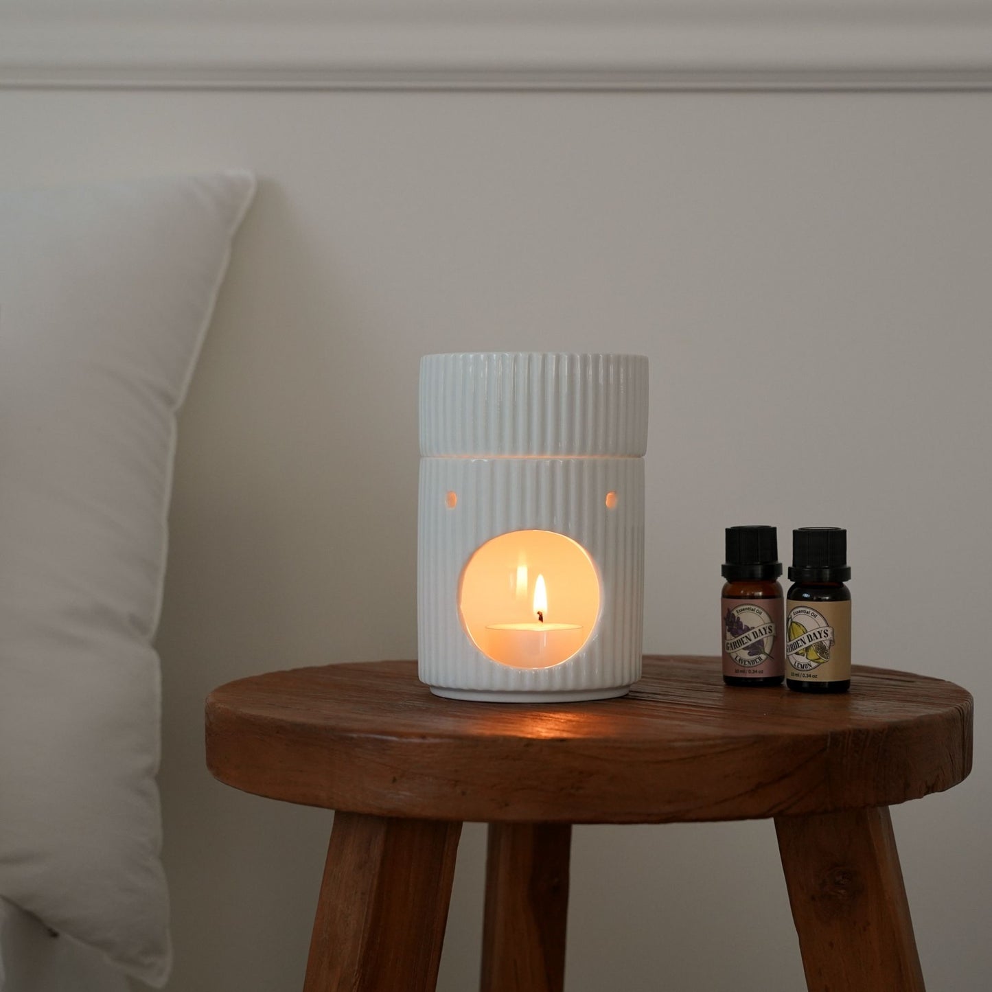 나무 스툴 위에서 티라이트 캔들이 켜진 화이트 라운드 오일버너와 옆에 세워둔 가든데이즈 에센셜오일 2병이 보이는 선물세트 사용 연출 사진