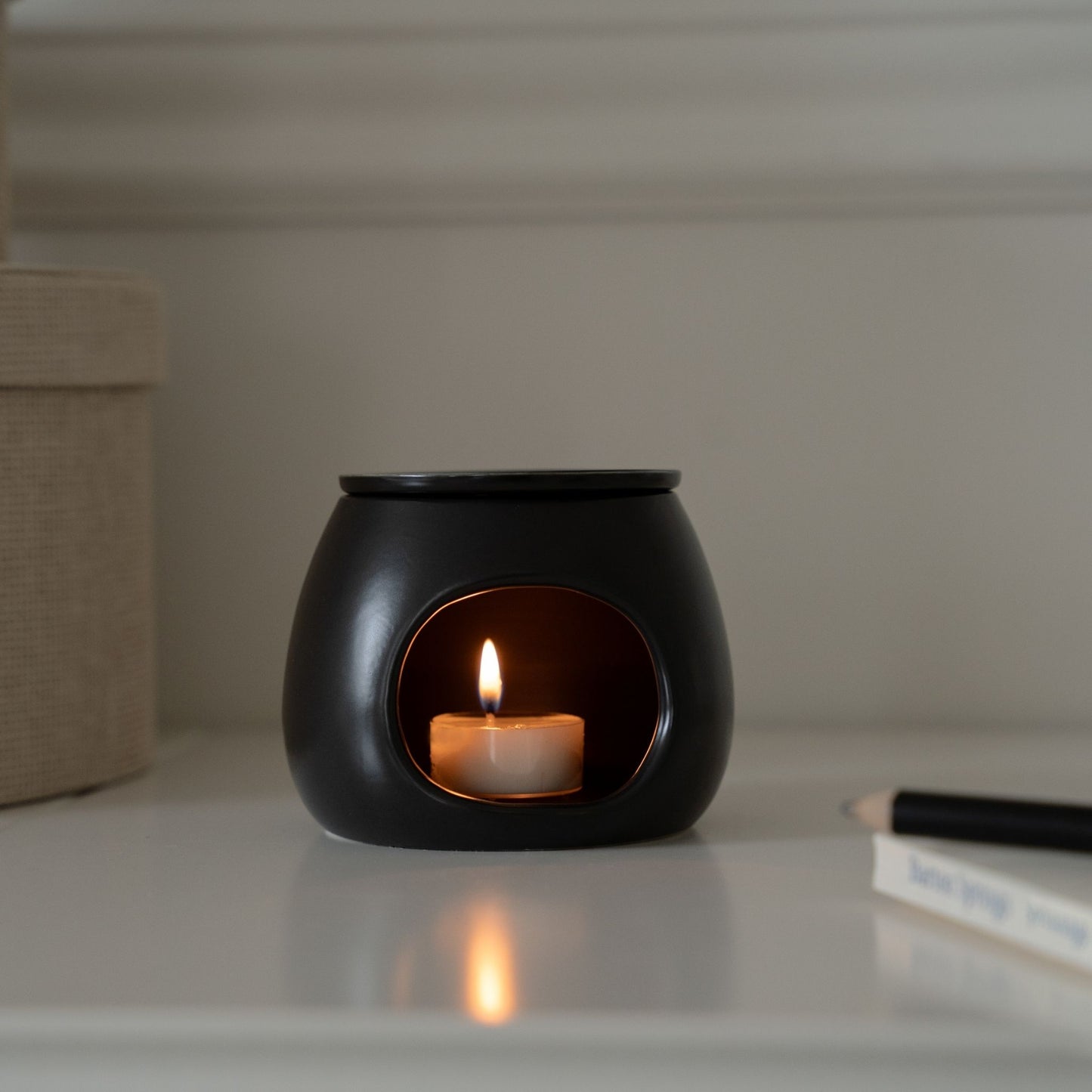 화이트 선반 위 책과 장식 오브제 옆에 놓인 블랙 세라믹 오일버너와 은은한 불빛의 티라이트 캔들이 보이는 인테리어 연출 사진