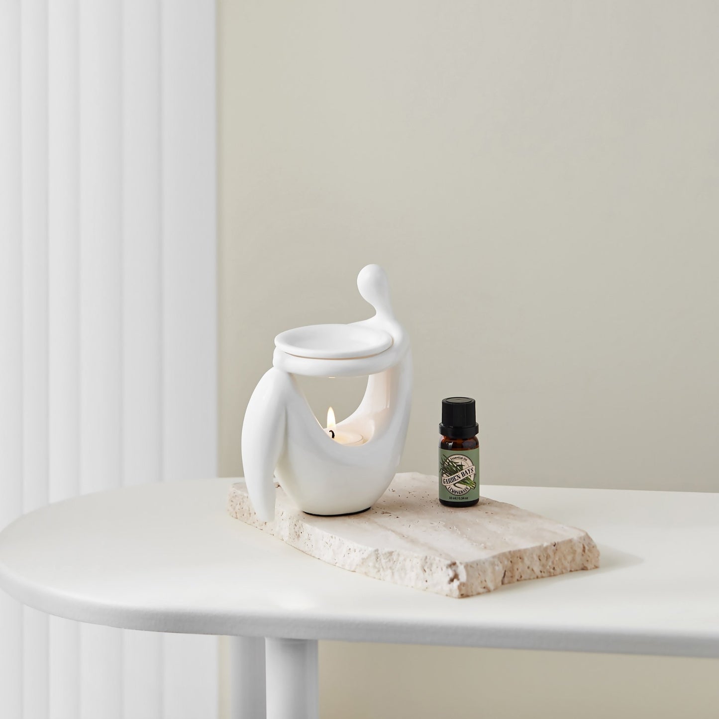 둥근 화이트 테이블 위 타월과 가든데이즈 에센셜오일 병 옆에 놓인 화이트 조형적인 세라믹 오일버너와 은은한 불빛의 티라이트 캔들이 보이는 연출 사진