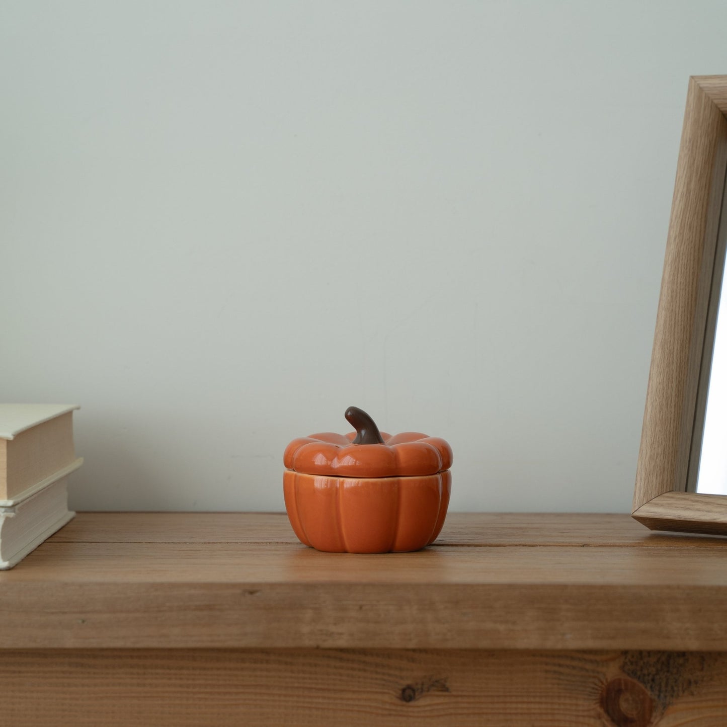 창가 쪽 나무 선반 위에 흰색 책과 함께 놓인 주황색 호박 캔들 홀더, 옆쪽에 살짝 보이는거울이 있는 연출 사진