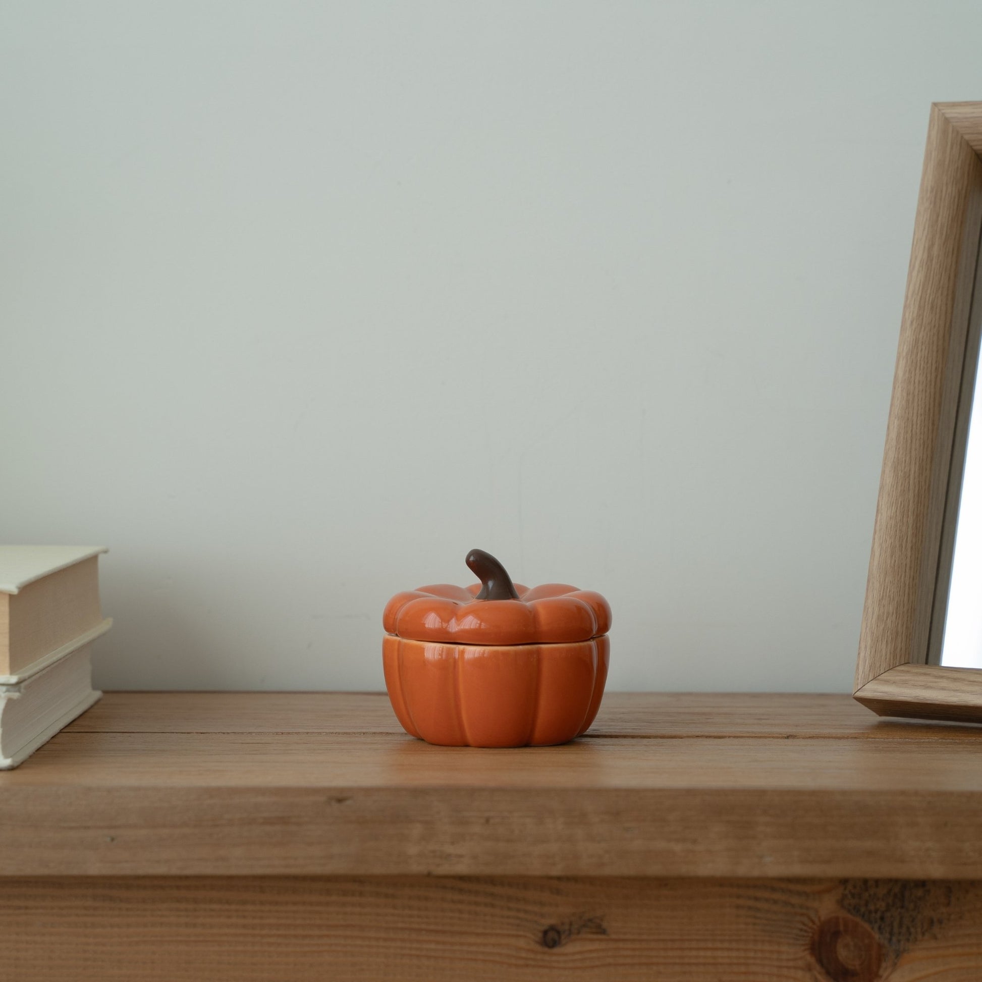 창가 쪽 나무 선반 위에 흰색 책과 함께 놓인 주황색 호박 캔들 홀더, 옆쪽에 살짝 보이는거울이 있는 연출 사진