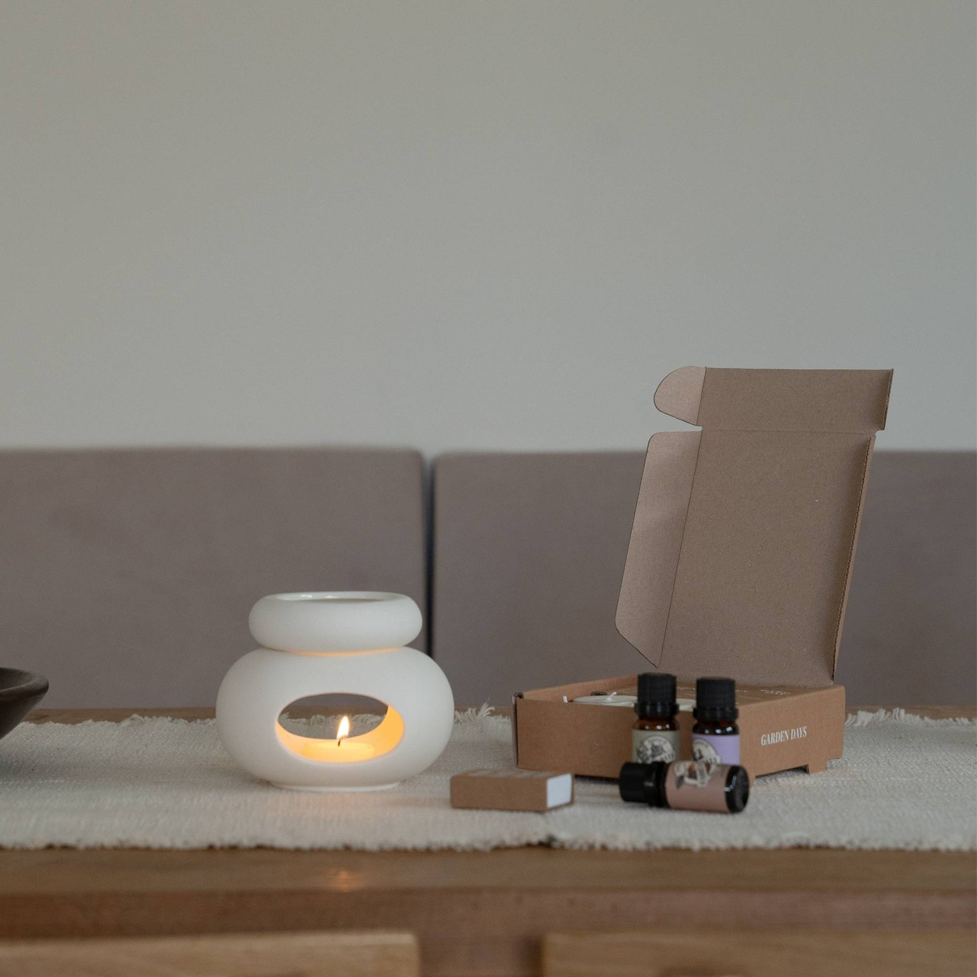  나무 테이블 위에서 티라이트 캔들이 켜진 조약돌 오일버너와 그 뒤에 세워둔 선물박스, 옆의 에센셜오일 병들이 보이는  연출 사진