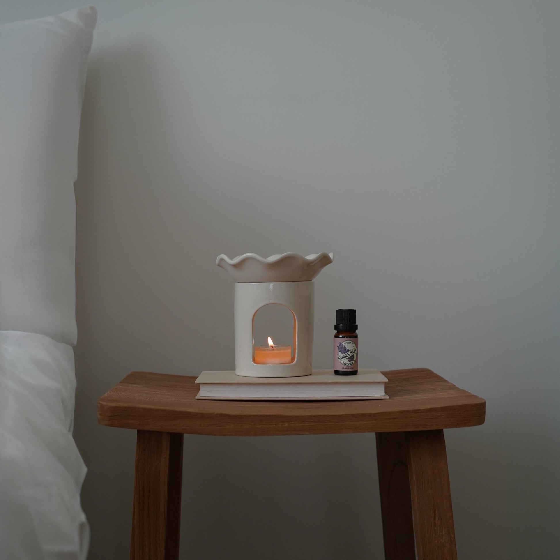 벽 앞 나무 벤치 위에 놓인 웨이브  모양 화이트 오일버너와 불이 켜진 티라이트 캔들, 옆에 세워둔 가든데이즈 에센셜오일 병 두 개가 보이는 연출 사진