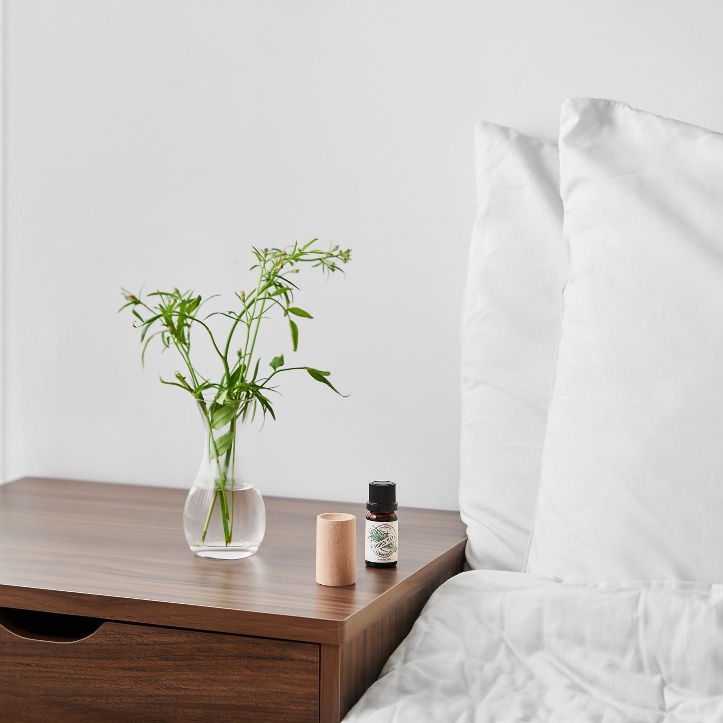 침대 옆 나무 협탁 위 유리 화병과 가든데이즈 에센셜오일 병 옆에 세워진 너도밤나무 원목 실린더 발향 우드가 보이는 침실 연출 사진