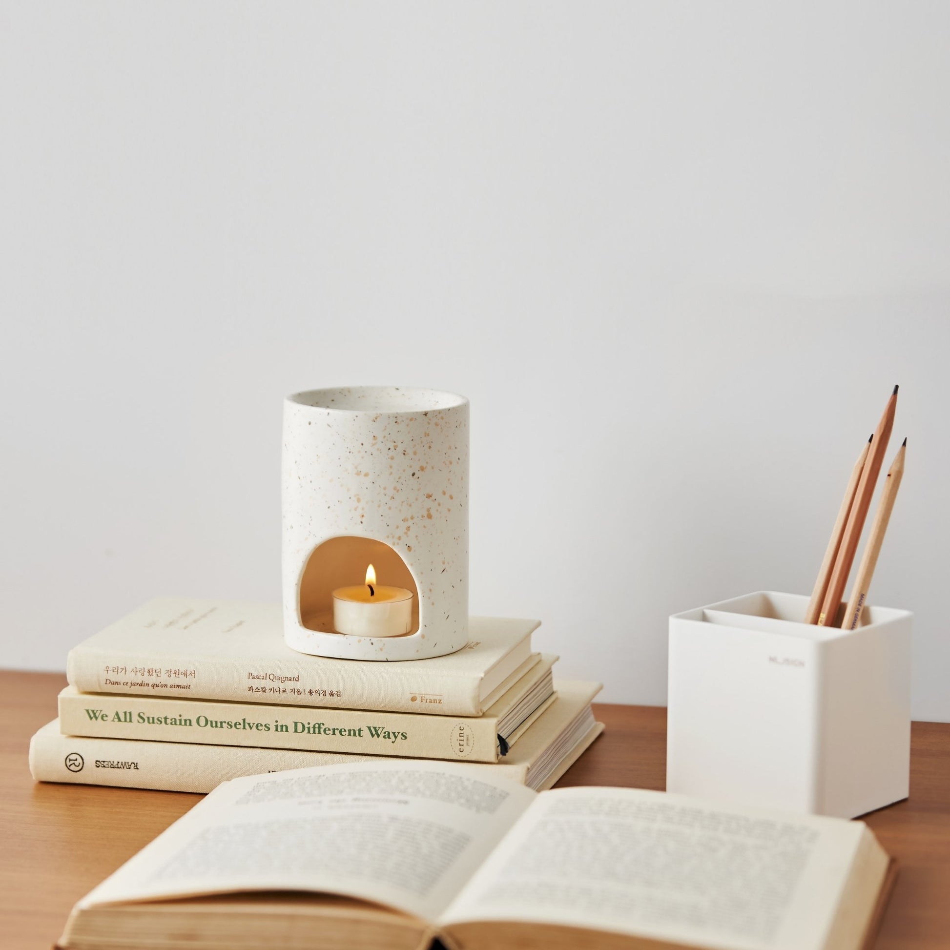 책이 쌓인 나무 책상 위에 올려진 마블 텍스처 오일버너와 따뜻한 불빛의 티라이트 캔들, 옆에 펜꽂이와 펼쳐진 책이 함께 보이는 작업 공간 연출 사진