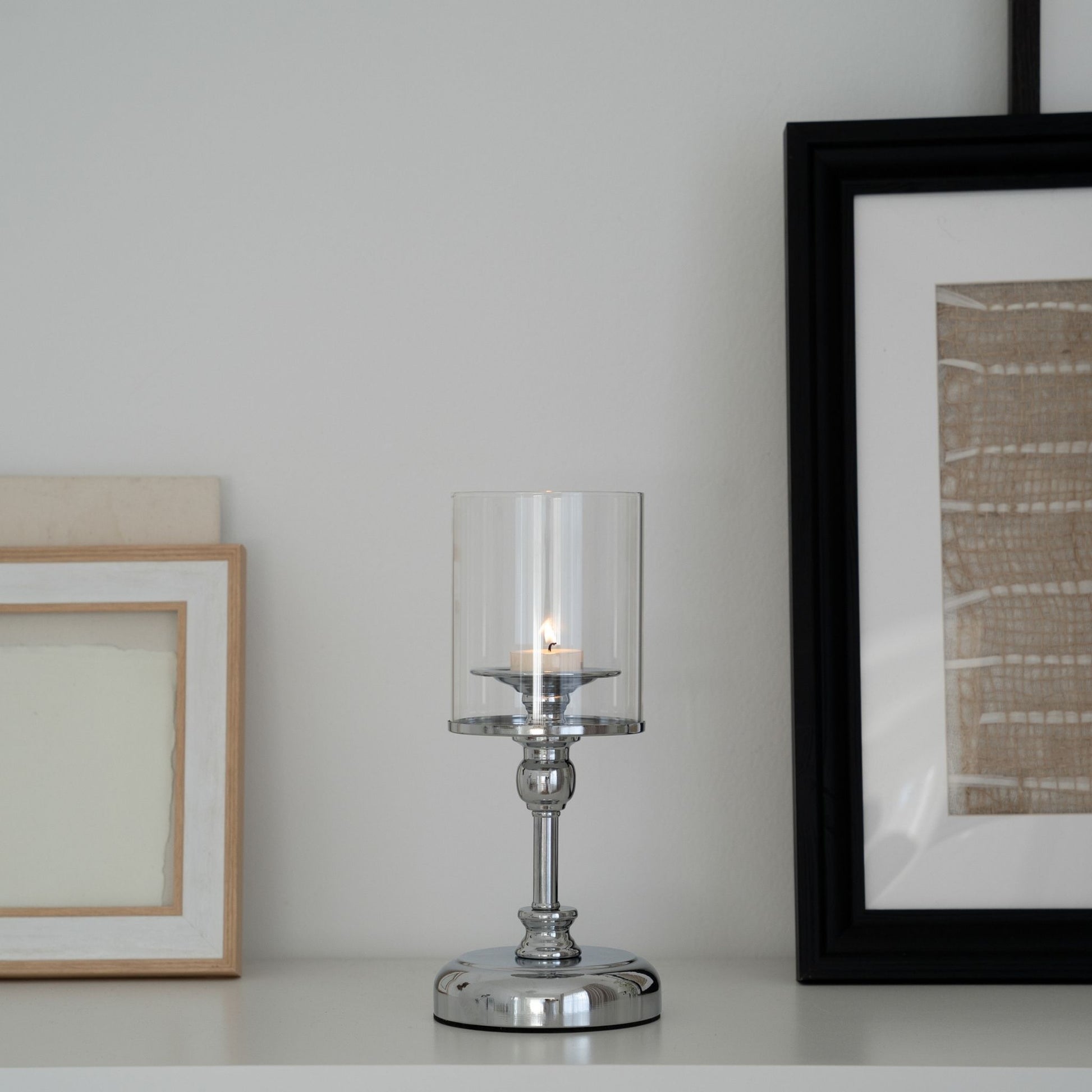 액자와 오브제가 놓인 선반 위에 실버 스탠드 유리 캔들 홀더 한 개와 안에서 은은하게 켜진 티라이트 캔들이 보이는 인테리어 연출 사진
