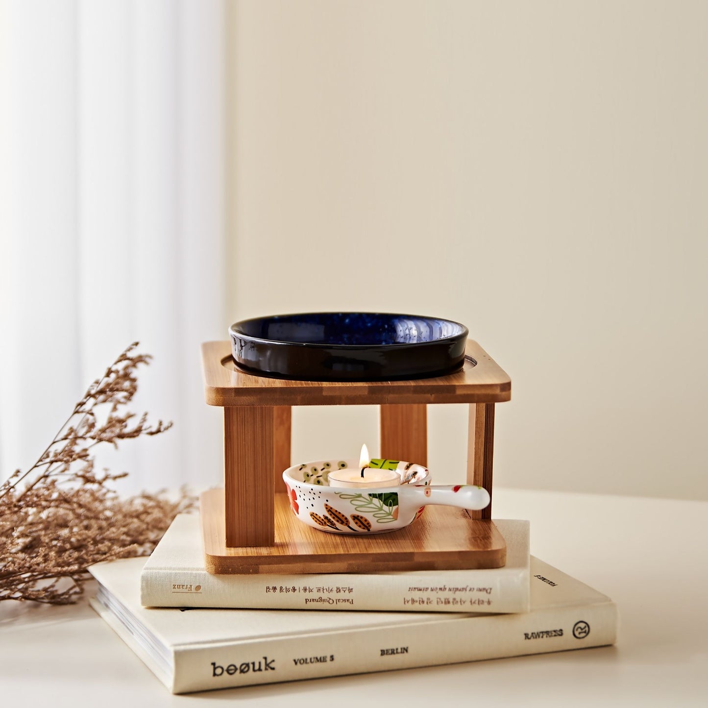 밝은 배경 앞 책 더미 위에 올려진 우드 스탠드 오일버너와 네이비 세라믹 볼, 아래 불이 켜진 티라이트 캔들, 옆에 말린 꽃 장식이 놓인 연출 사진