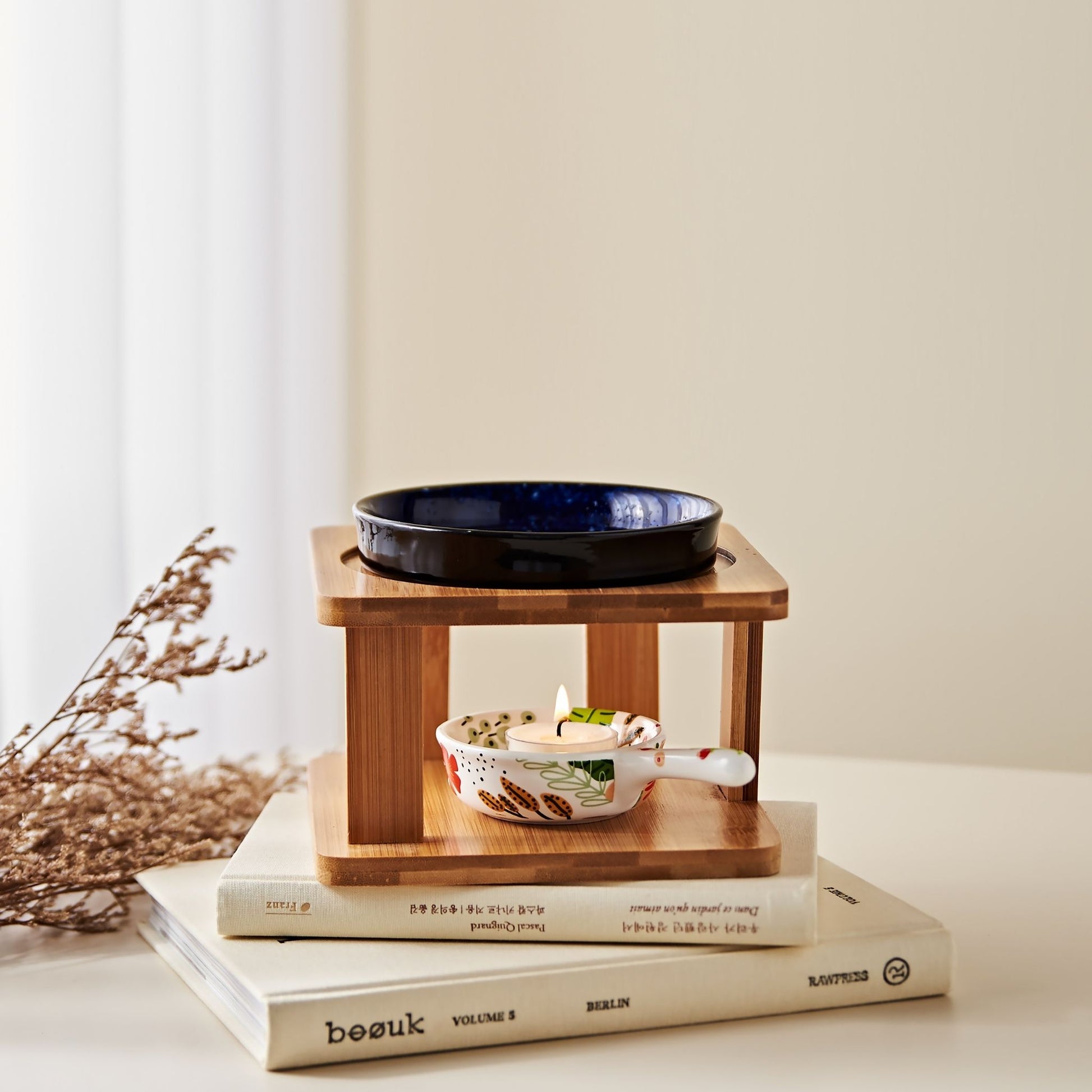 밝은 배경 앞 책 더미 위에 올려진 우드 스탠드 오일버너와 네이비 세라믹 볼, 아래 불이 켜진 티라이트 캔들, 옆에 말린 꽃 장식이 놓인 연출 사진