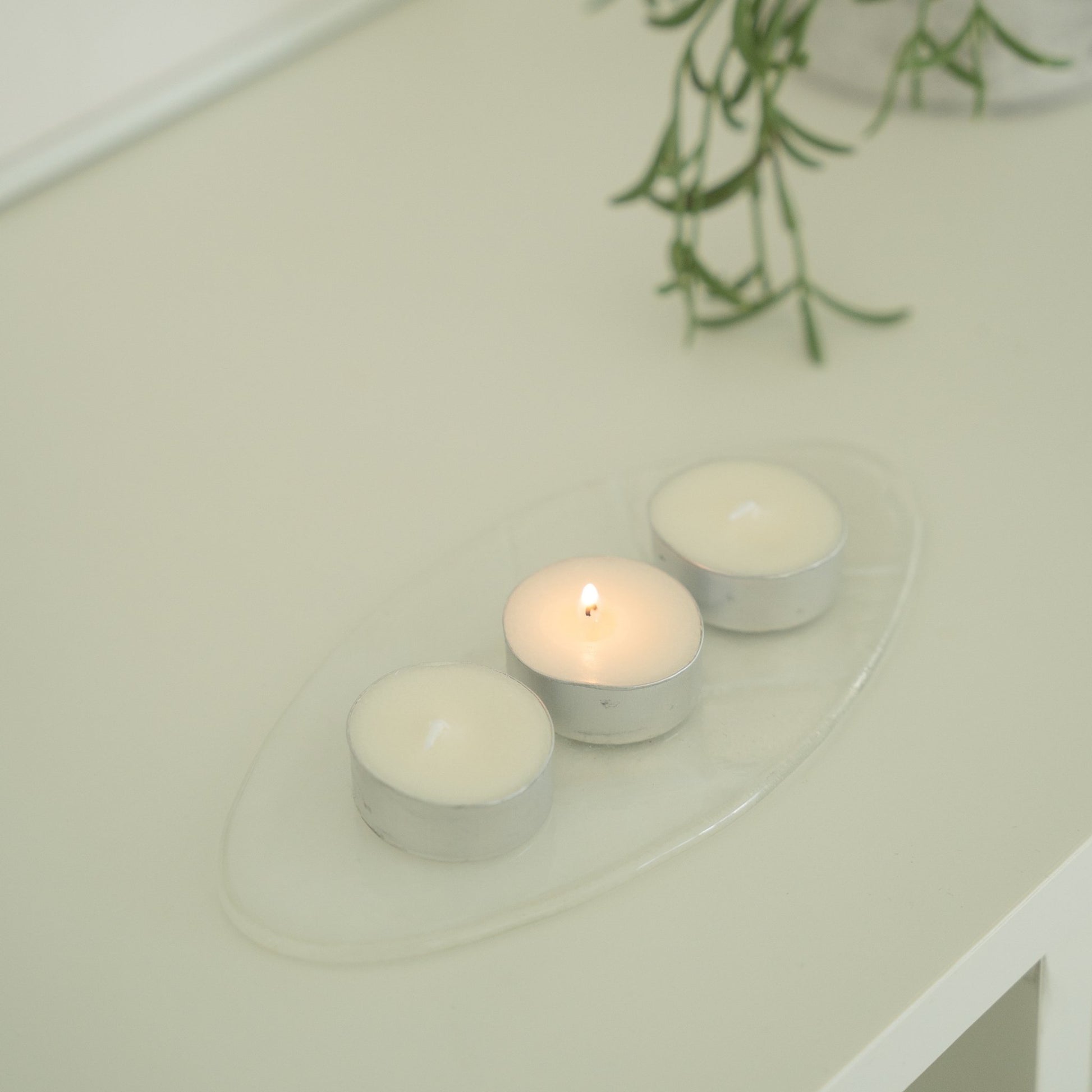 초록 식물이 드리운 화이트 상판 위 투명 받침 접시 위에 올려진 티라이트 캔들 세 개가 보이는 제품 연출 사진