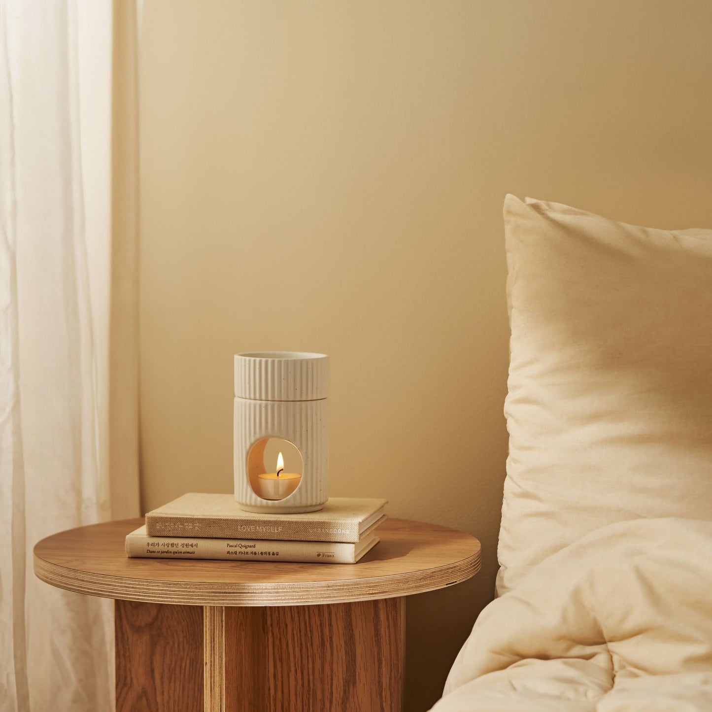 침대 옆 둥근 나무 협탁 위에 놓인 세로 골 패턴 도자기 오일버너와 불이 켜진 티라이트 캔들, 뒤로 커튼과 침구가 보이는 침실 연출 사진