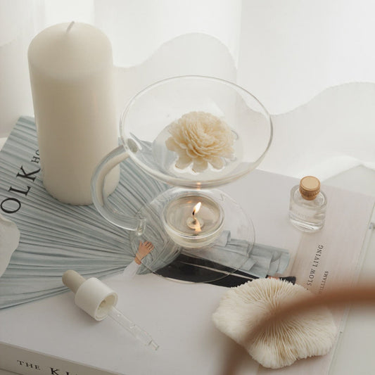 창가 앞 책 더미 위에 놓인 투명한 유리 오일버너와 아래에서 은은하게 빛나는 티라이트 캔들, 위에 올려진 플라워 디퓨저와 옆의 기둥 캔들과 장식이 함께 보이는 연출 사진