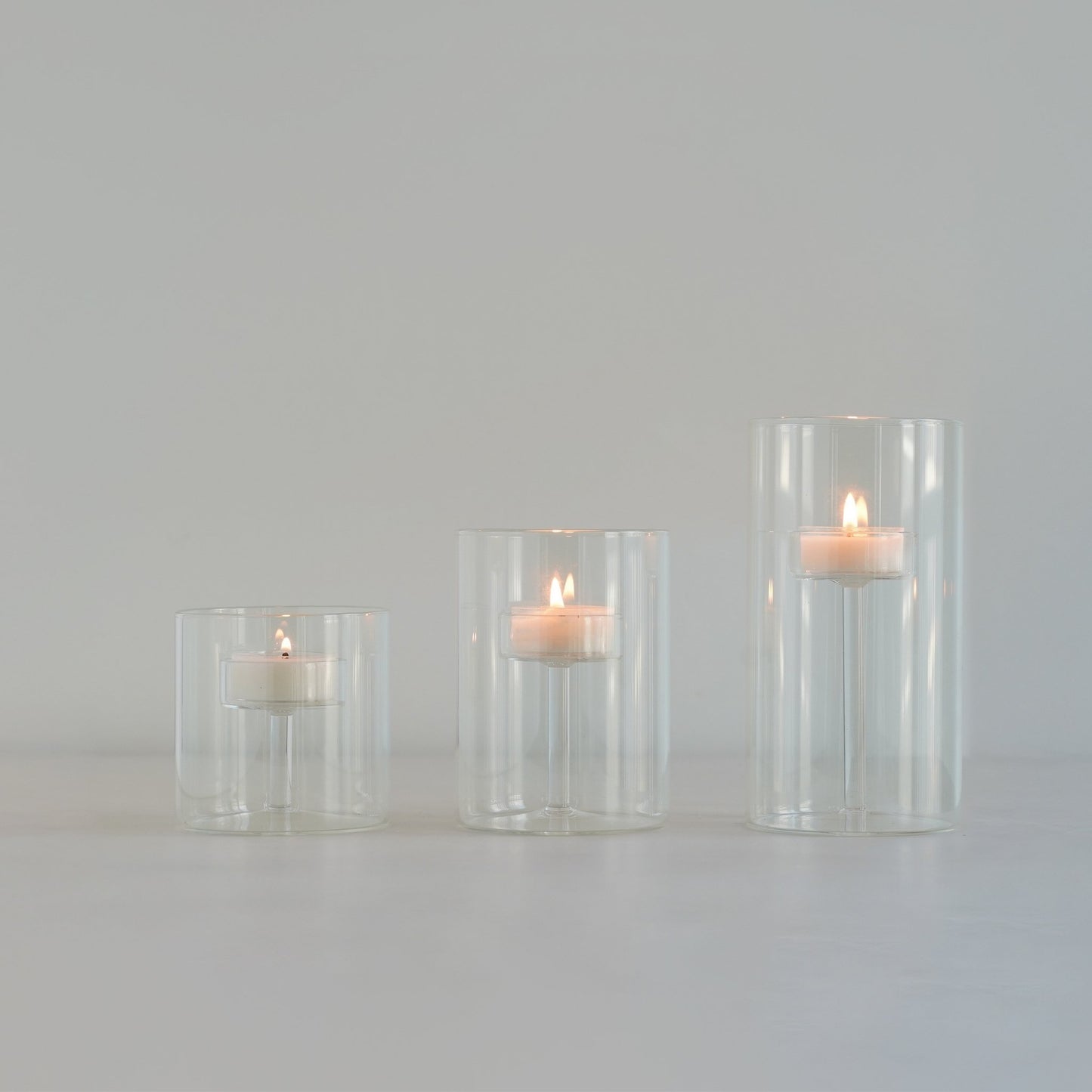 연한 배경 위에 줄지어 놓인 투명 유리 실린더 캔들 홀더와 안에서 따뜻하게 켜진 티라이트 캔들이 보이는 미니멀 제품 연출 사진
